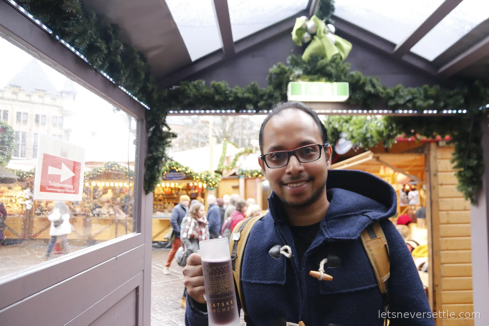 Dileep smiling under a garlanded market walkway with a Ratskeller Aachen mug of Gluhwein, stalls and the old town behind