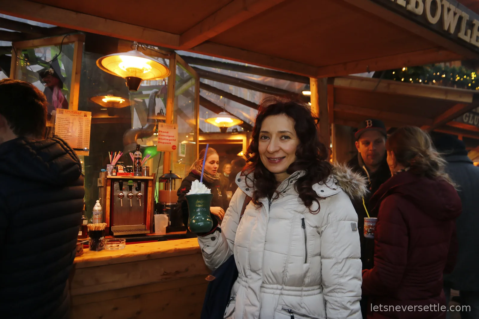 Inga in a white winter coat holding a green Aachen mug topped with whipped cream at the Hexenhof stall, a menu of Gluhwein variants behind