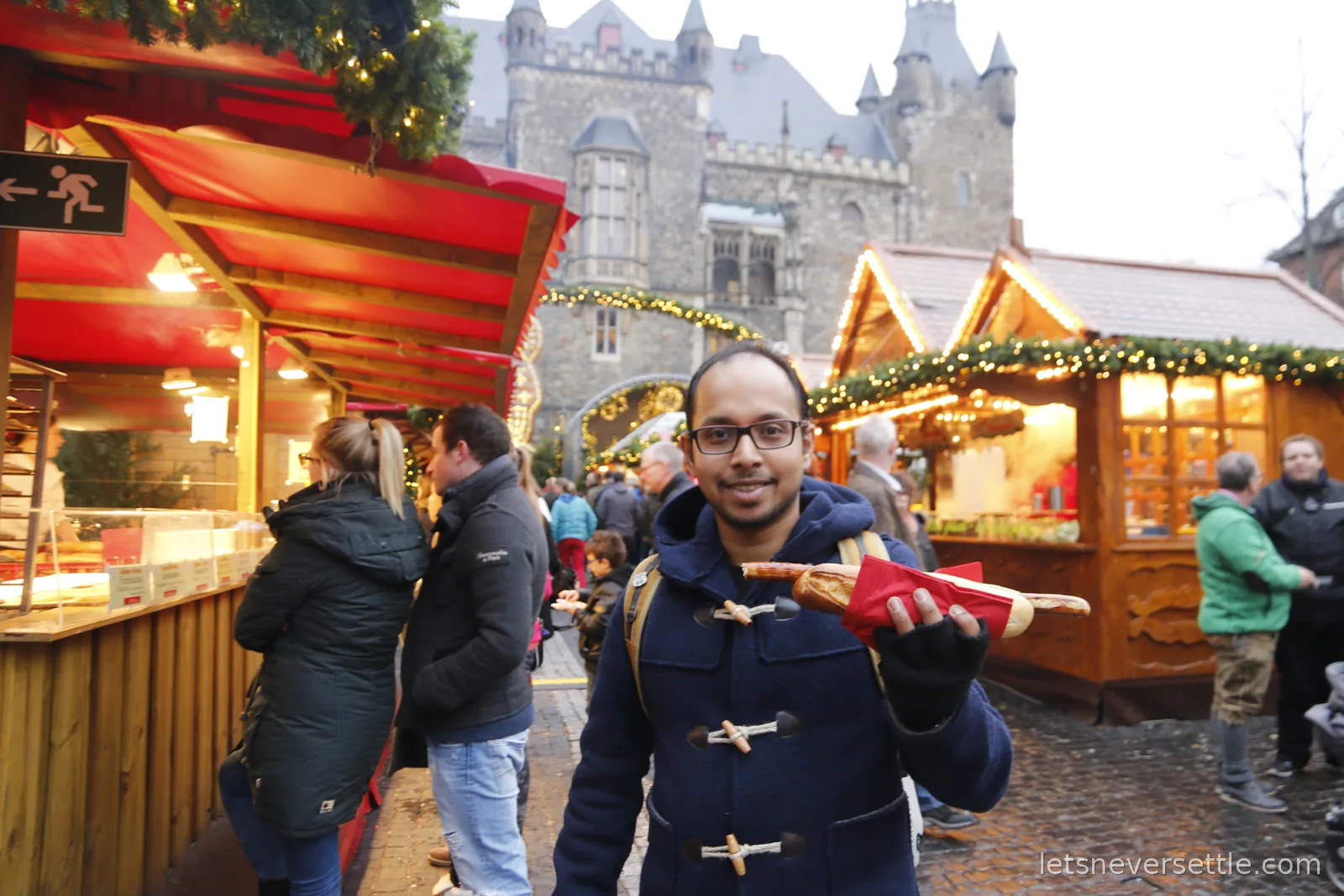 Dileep in a duffle coat holding a long bratwurst in a short baguette, wooden market chalets and the gothic Aachen Rathaus behind