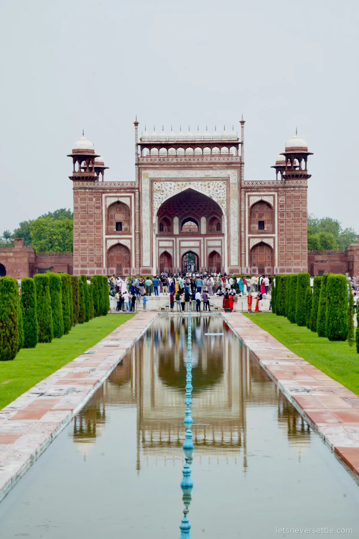 The red sandstone Great Gate of the Taj Mahal complex reflected in a long pool lined with cypress hedges, crowds passing under its central arch