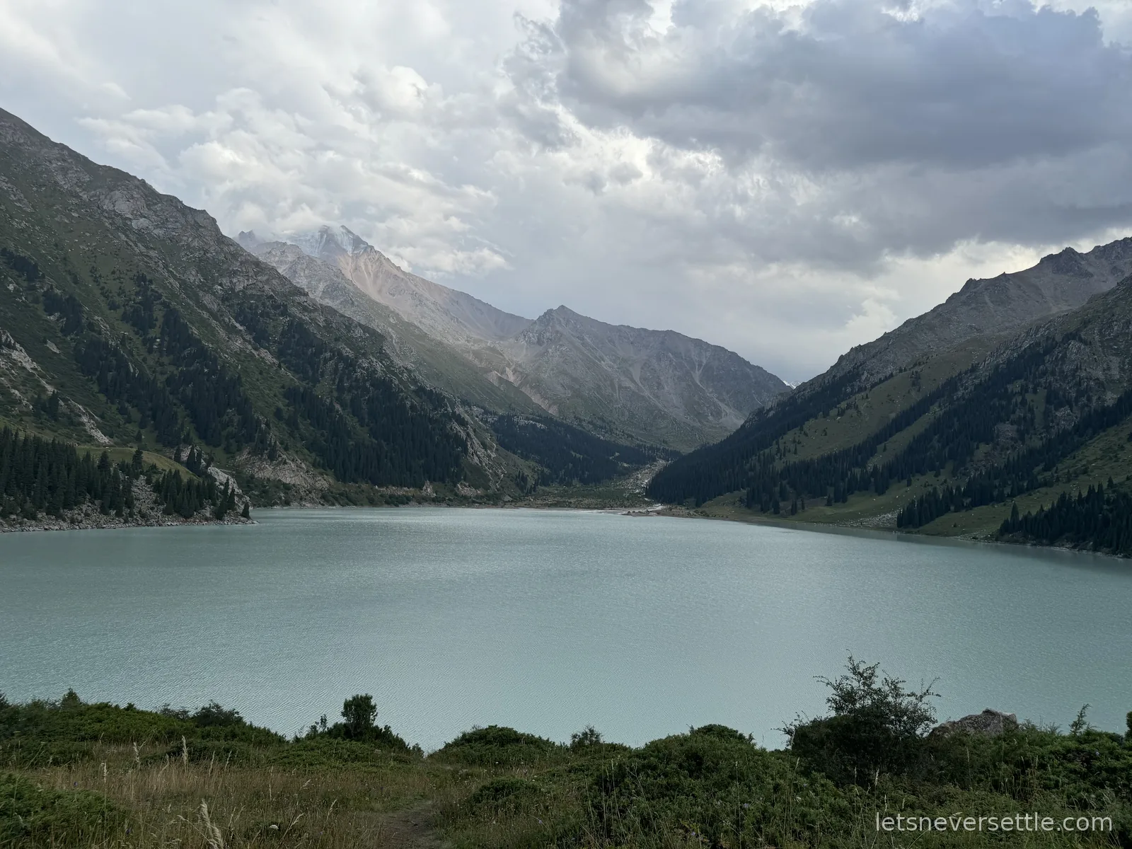 Big Almaty Lake under heavy clouds, pale glacial water with forested slopes and bare peaks rising on both sides