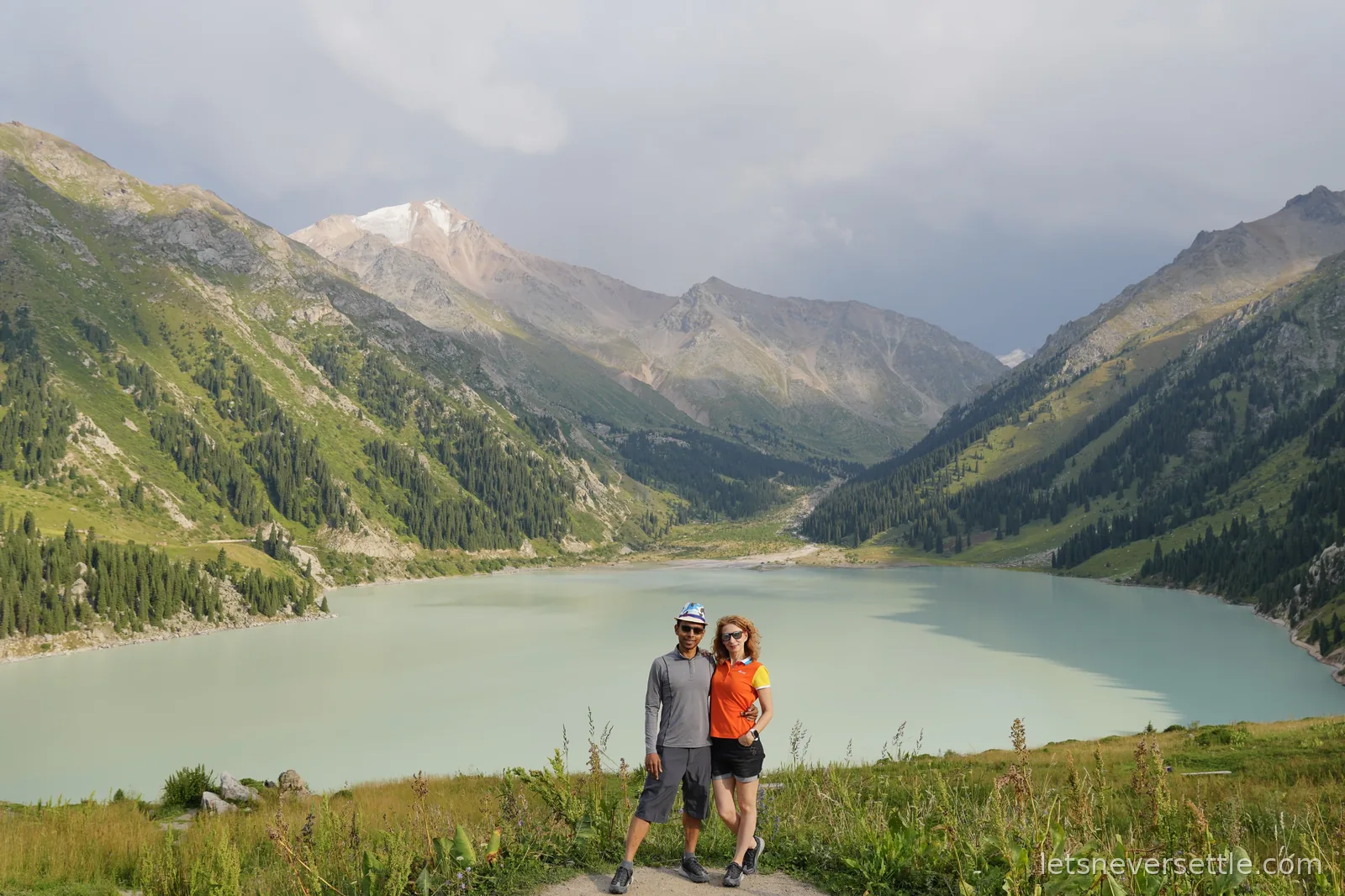 Inga and Dileep standing on a grassy viewpoint above Big Almaty Lake, its pale turquoise water ringed by fir-covered mountains