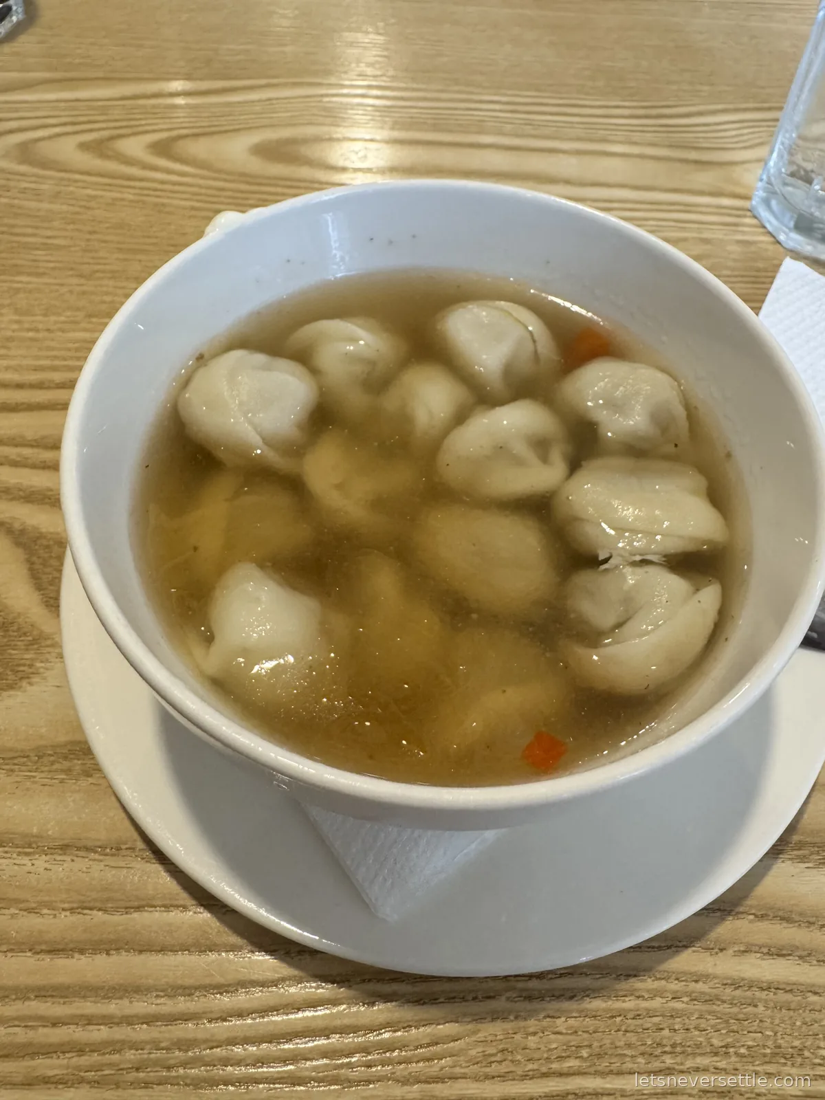 A white bowl of dumplings in clear broth with a cube of carrot, on a pale wooden table