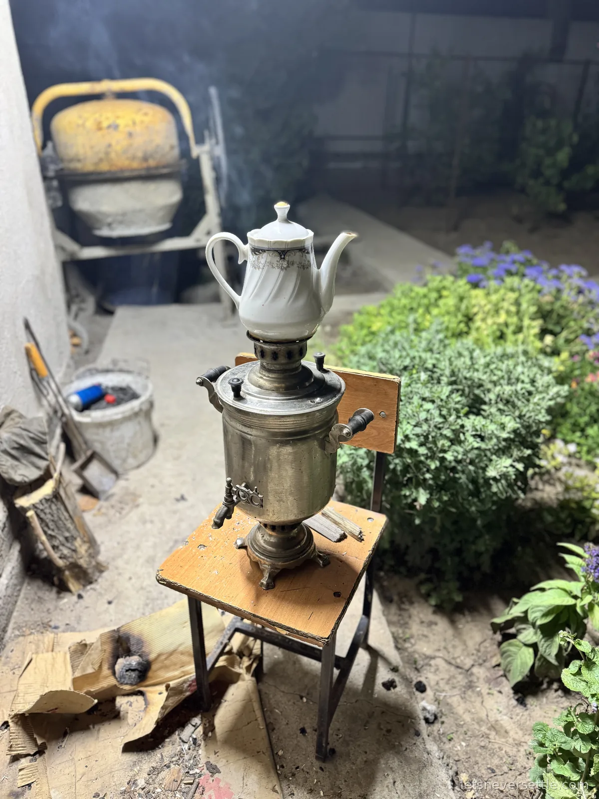 A wood-fired metal samovar smoking on a stool in a garden at night, a white porcelain teapot warming on its chimney