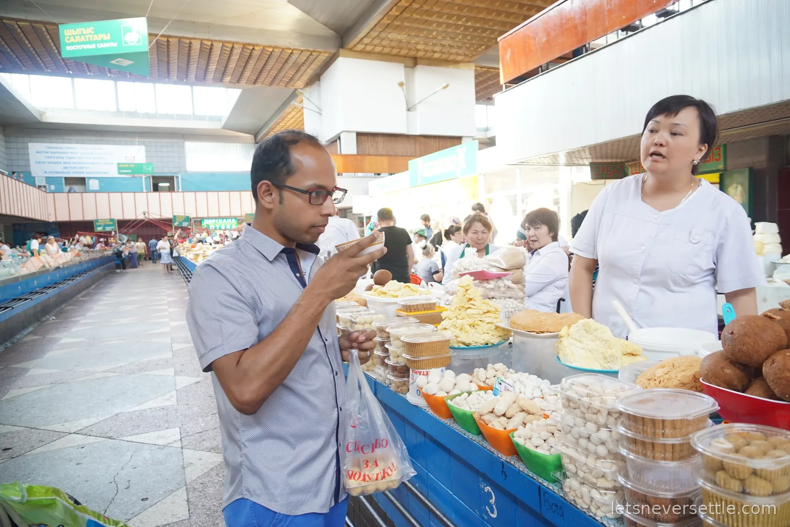 Dileep sampling from a small cup at a dairy counter in the Green Bazaar, tubs of white kurt balls and mounds of cheese along the stall