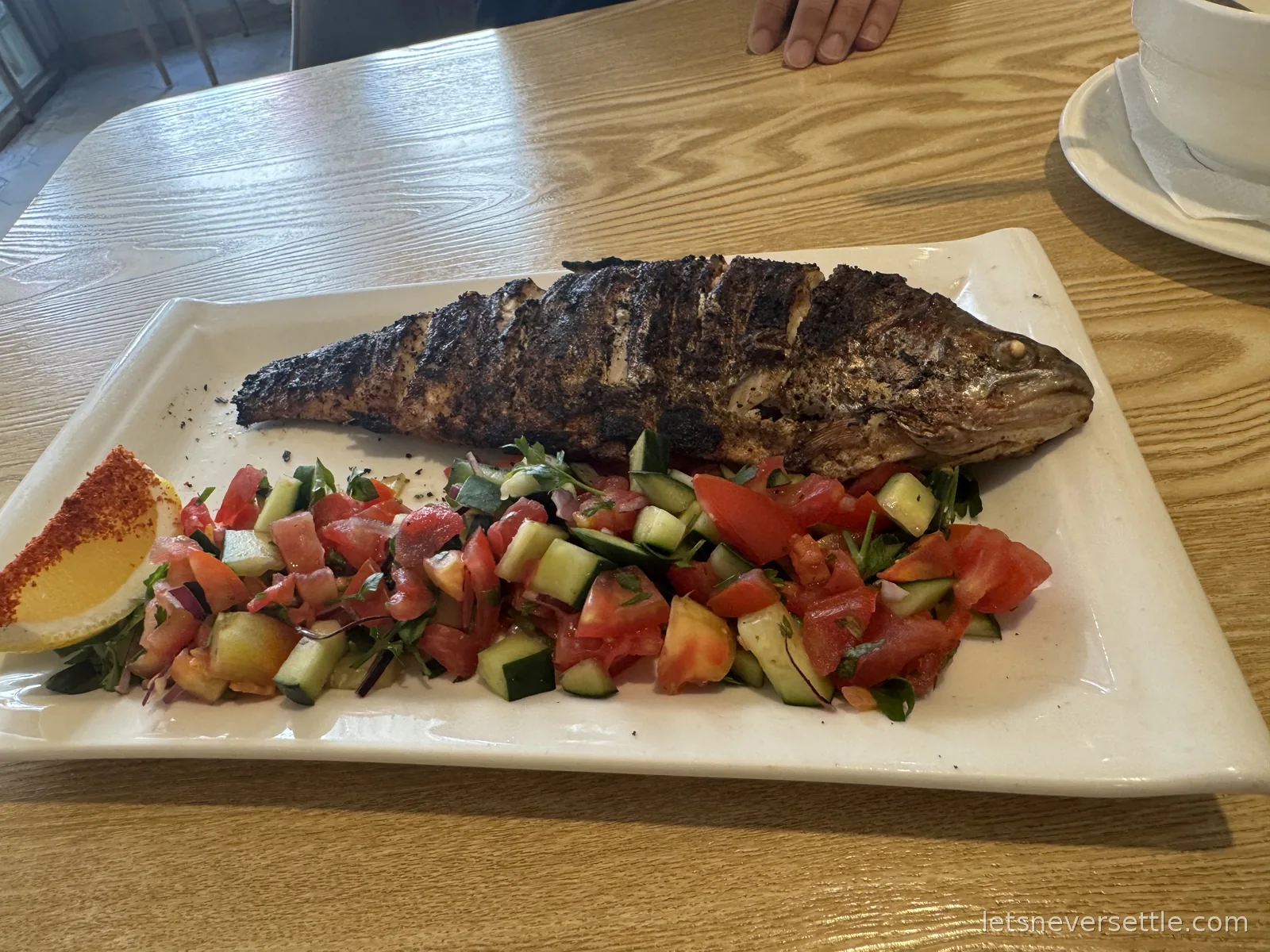 A whole grilled fish on a white platter with chopped tomato and cucumber salad and a spiced lemon wedge
