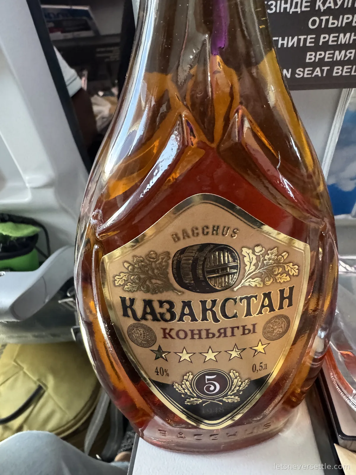 A bottle of Bacchus Kazakhstan cognac with a five star label standing on an airline seat tray, a Kazakh-language seat belt notice behind it