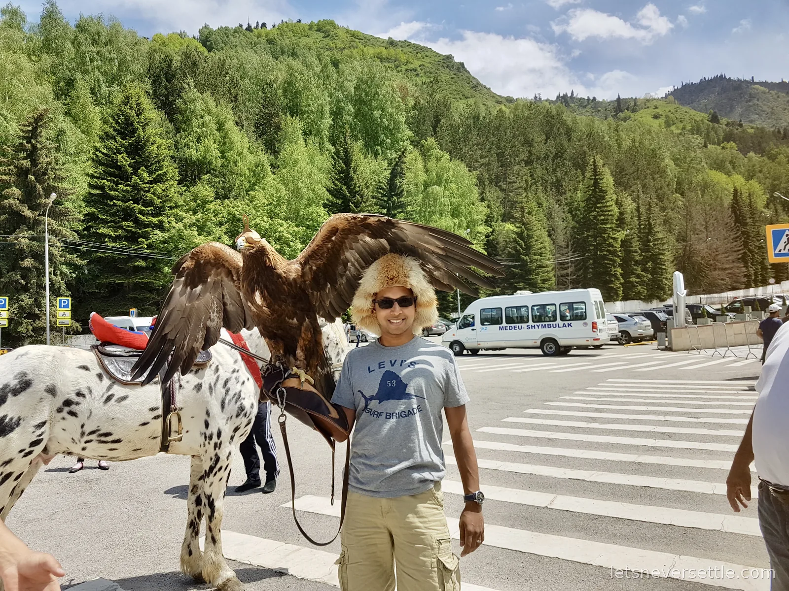 Dileep in a fur hat and sunglasses with a golden eagle spreading its wings above his shoulder, a spotted horse beside him and forested hills behind