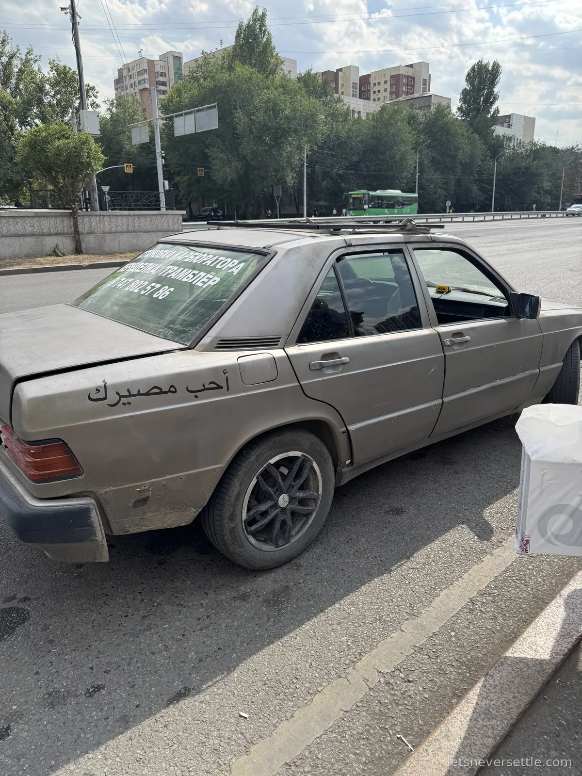 An old beige Mercedes saloon parked on an Almaty street with Arabic calligraphy painted along its side