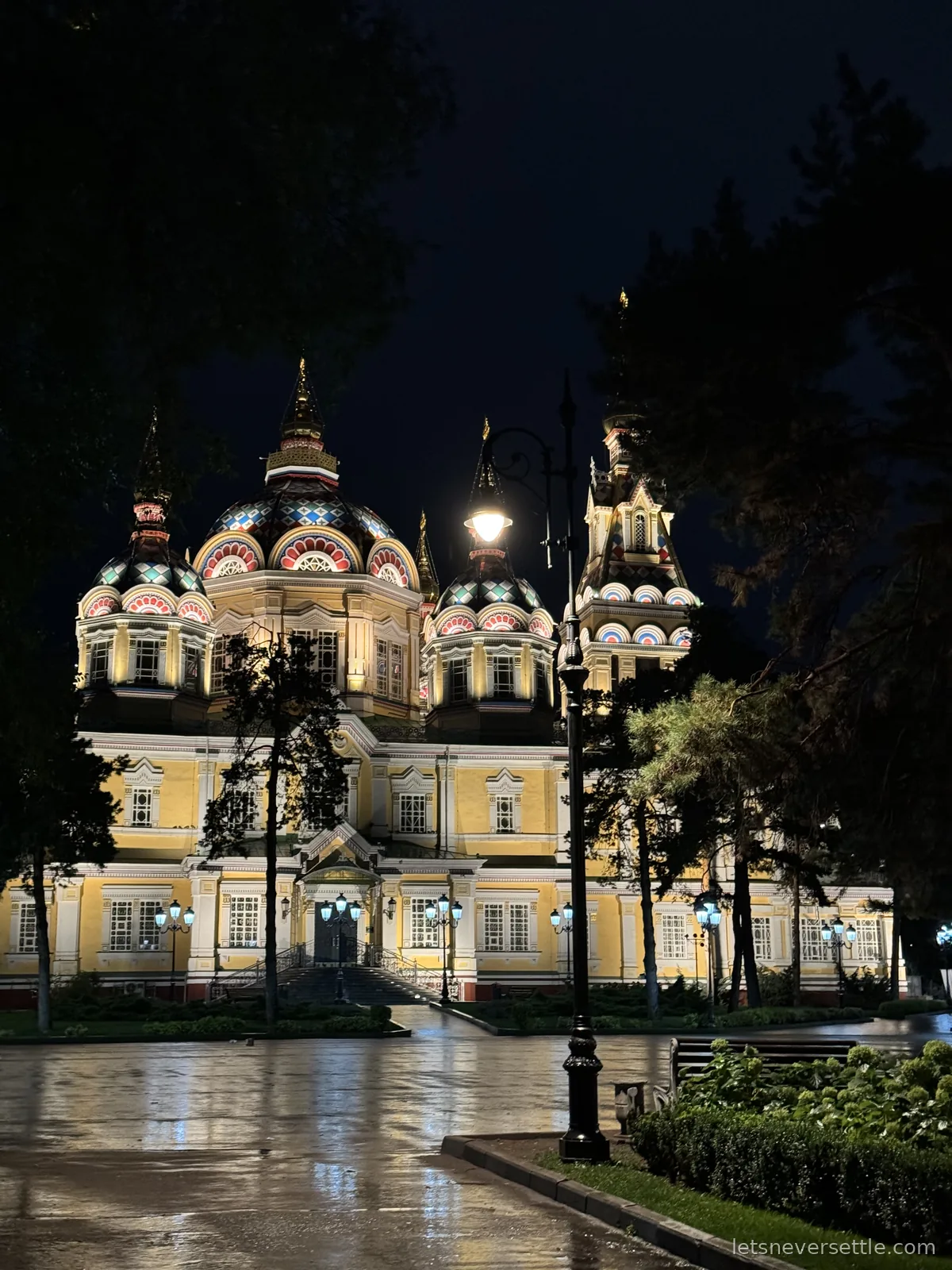 The cathedral lit up at night, painted domes and yellow walls glowing above wet paving and park lamps
