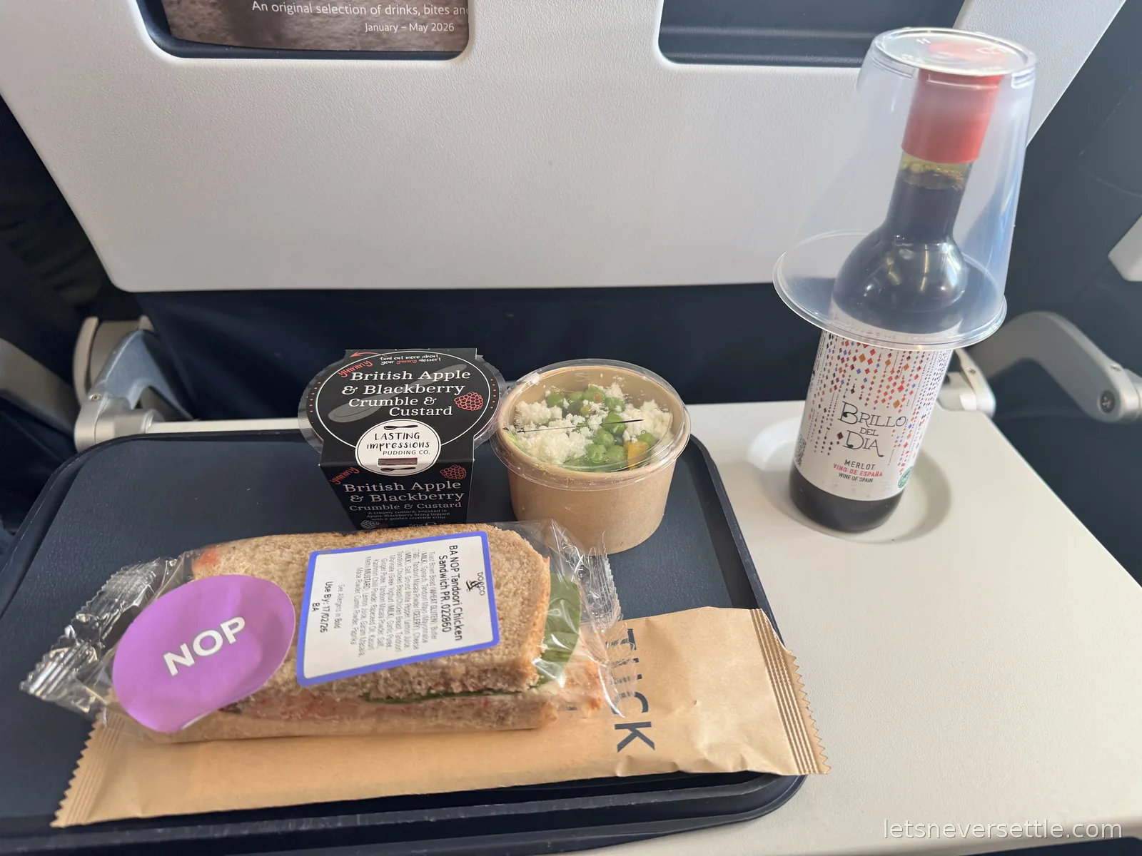 A British Airways economy meal tray with a wrapped sandwich, a salad pot, an apple and blackberry crumble and a small bottle of red wine