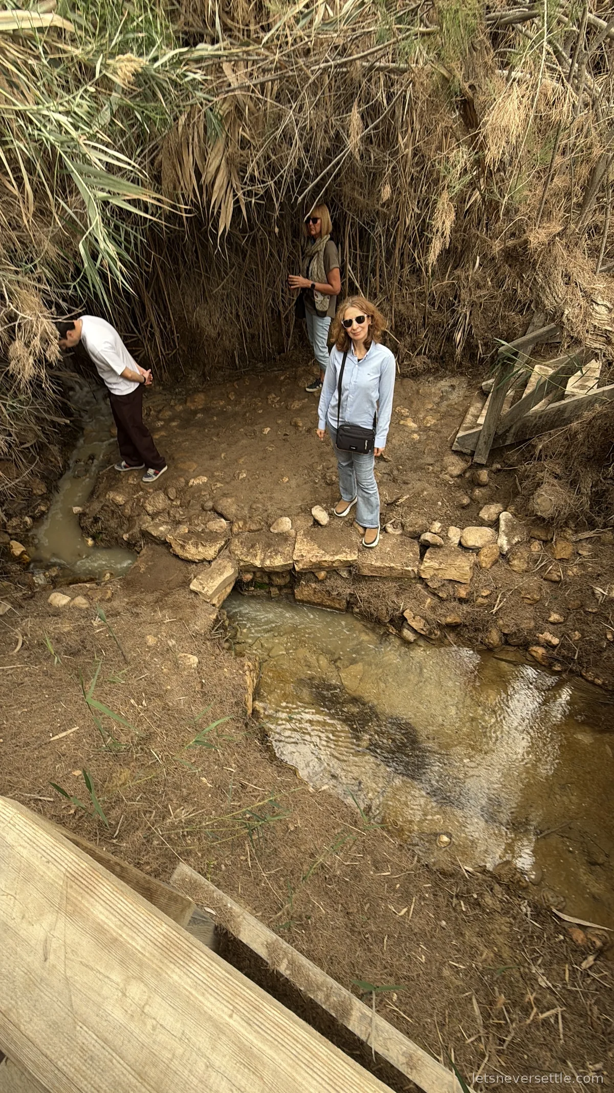 Inga standing on stones beside the shallow spring at Bethany Beyond the Jordan, surrounded by tall dry reeds