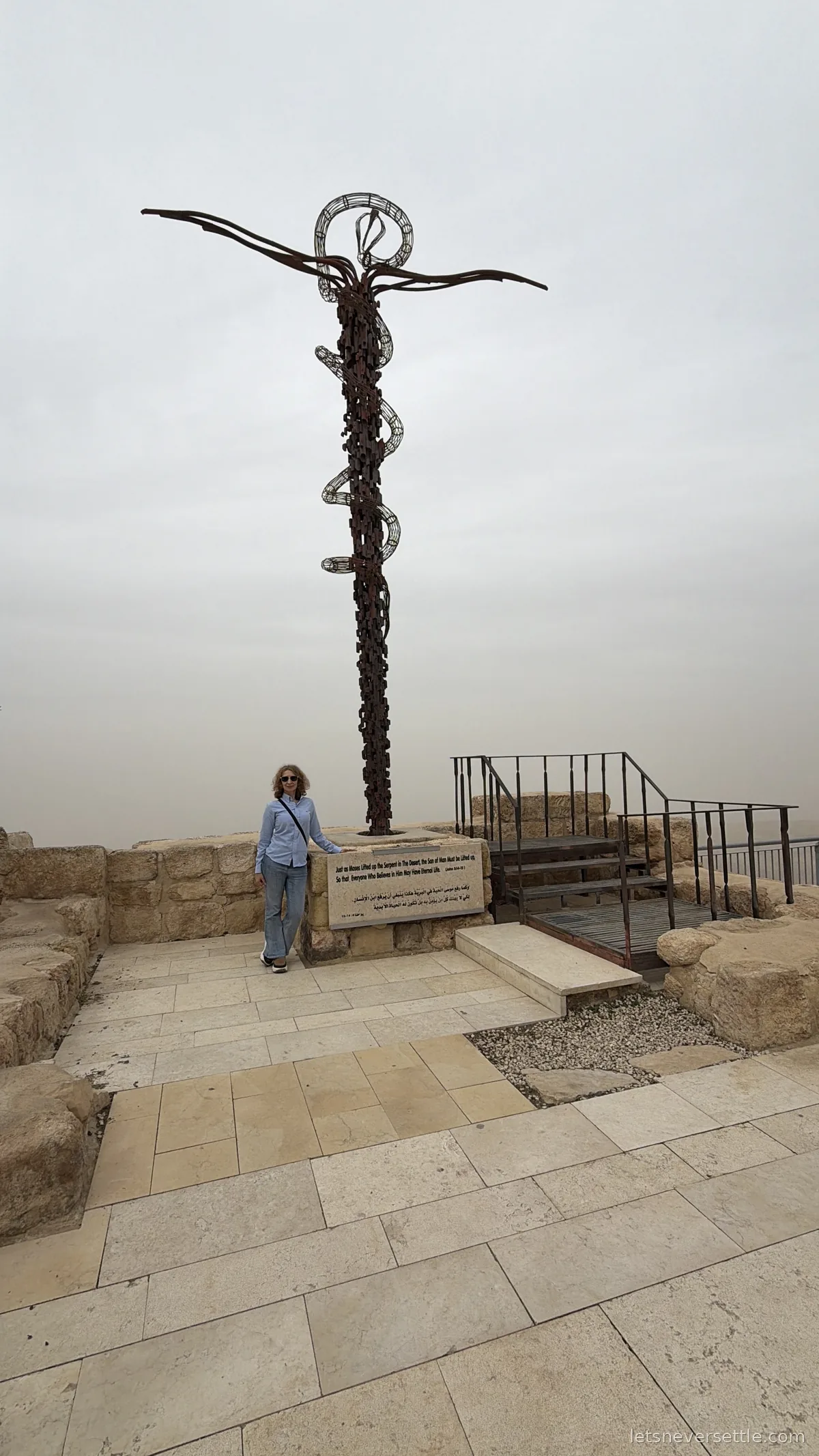 Inga standing beside the tall Brazen Serpent sculpture on Mount Nebo, a metal serpent coiled around a cross against a white sky