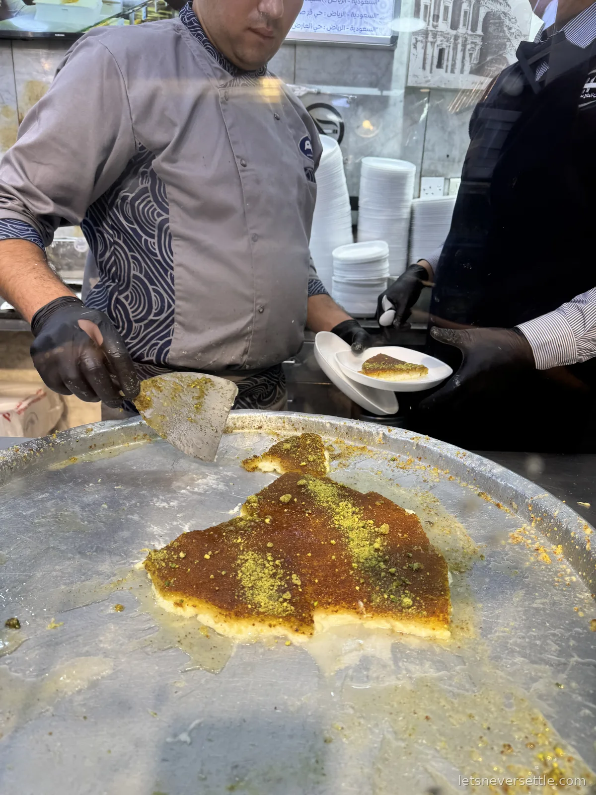 A chef lifting a slice of pistachio-topped kunafa off a huge round tray onto a plate at a sweet shop in Amman