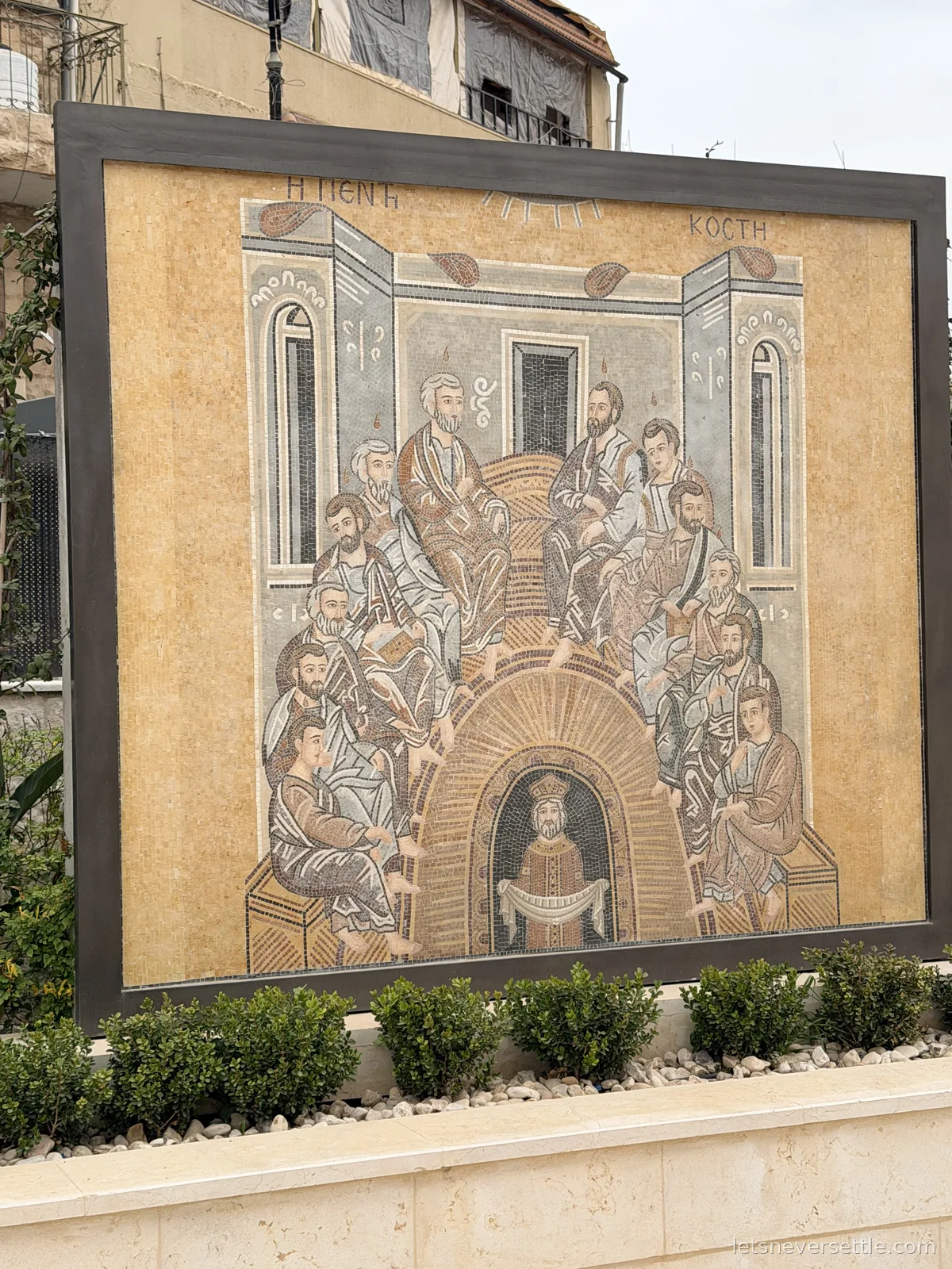 A large framed mosaic panel outdoors in Madaba showing the apostles seated in a semicircle, worked in pale stone tiles