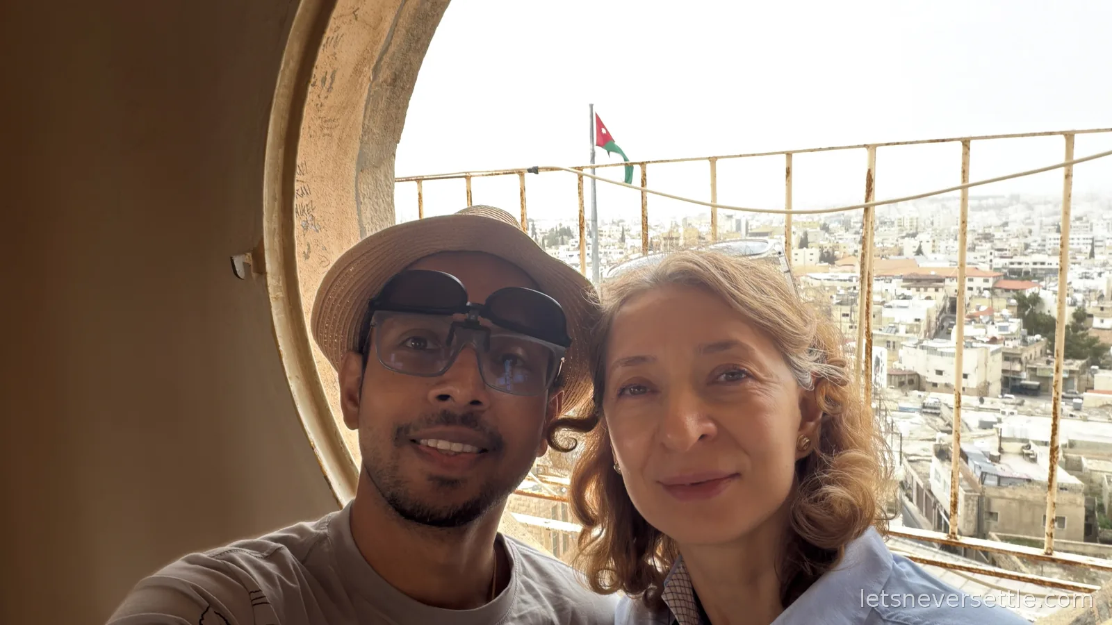 Inga and Dileep taking a selfie from a round window at the top of the bell tower in Madaba, a Jordanian flag flying above the town's rooftops behind them