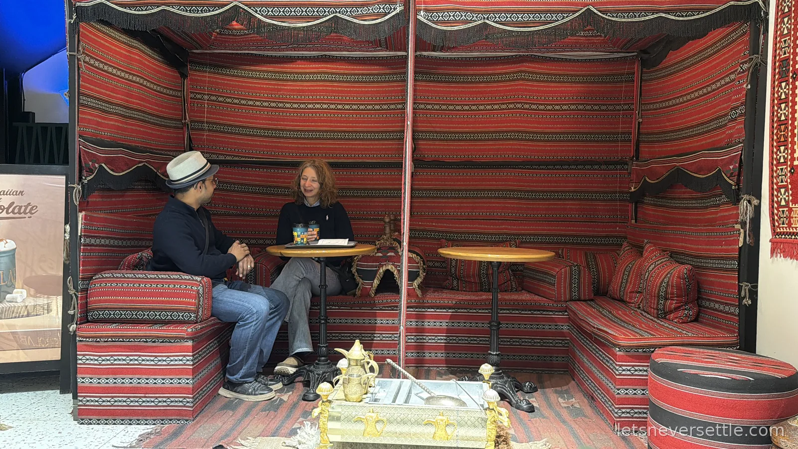 Inga and Dileep sitting in a Bedouin-style booth in Amman lined with red and black striped cushions, a brass coffee set on a tray in front of them