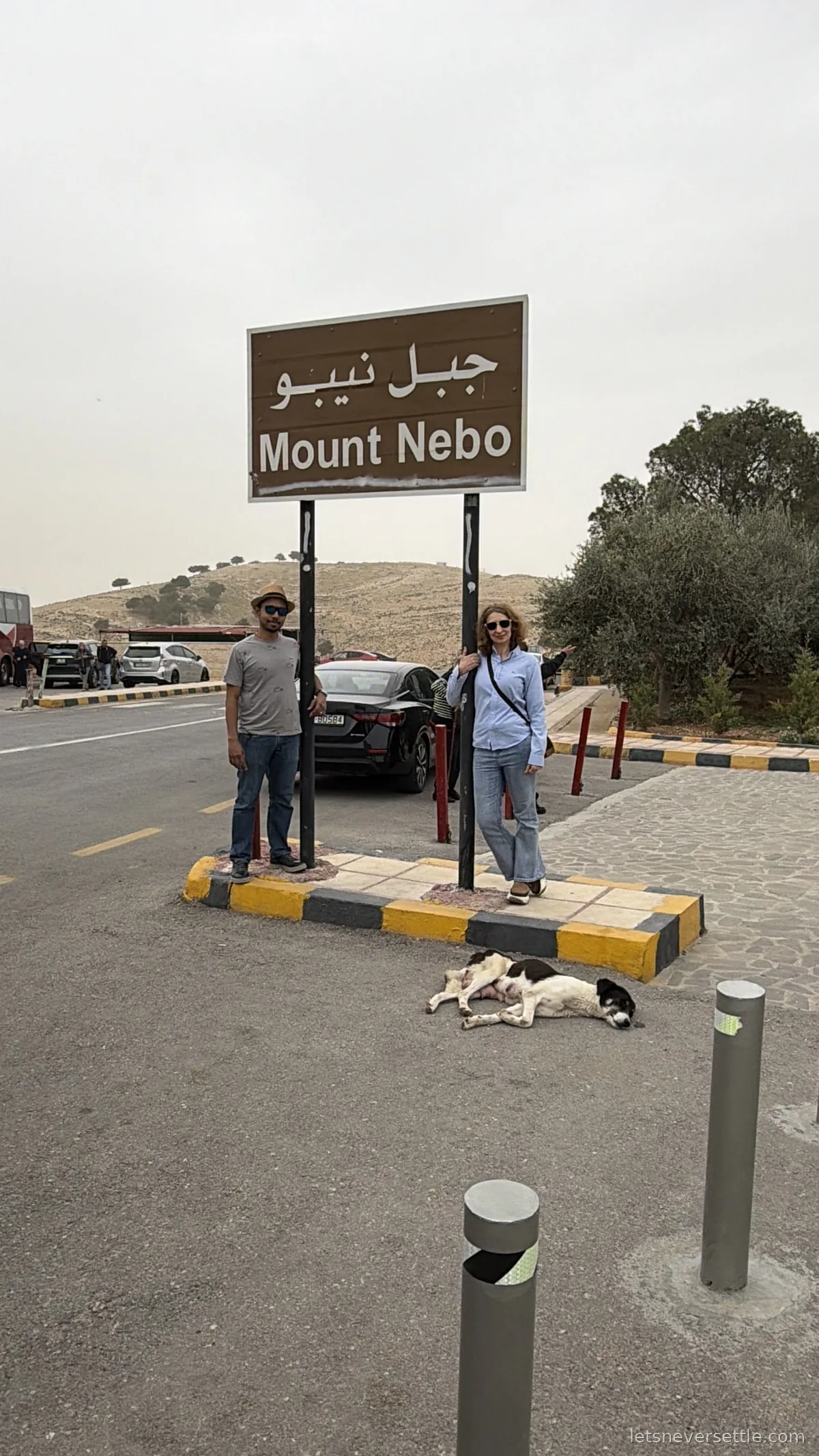 Inga and Dileep standing either side of a brown road sign reading Mount Nebo in English and Arabic, with a dog asleep on the kerb below it