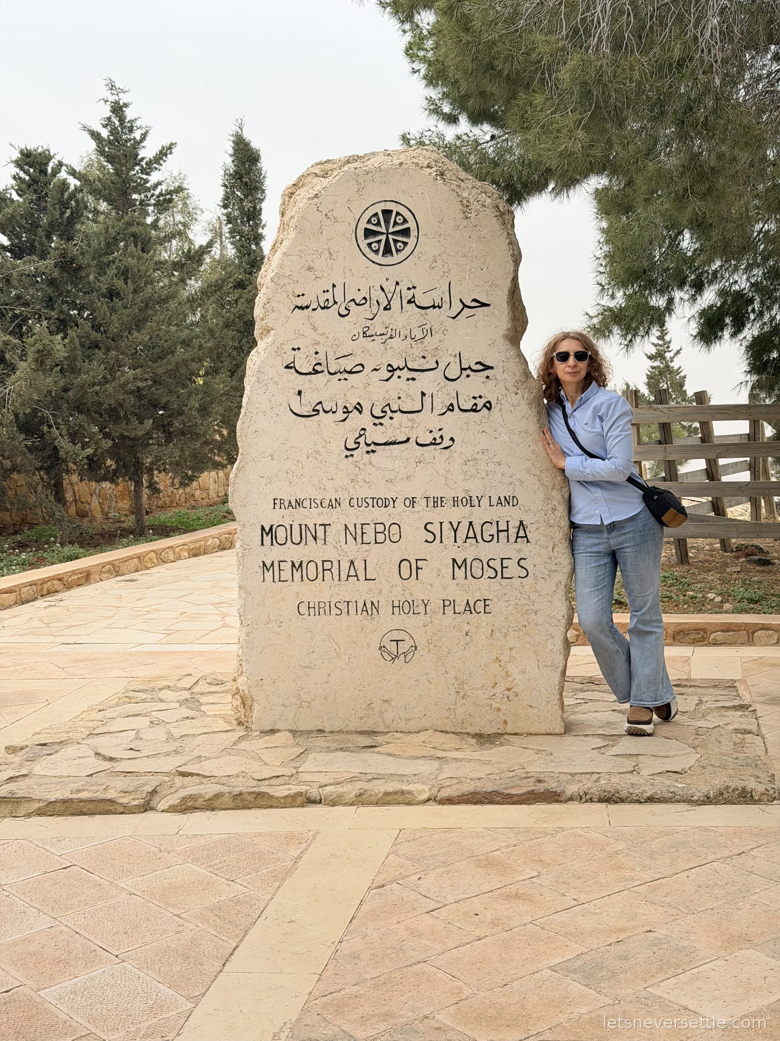 Inga leaning on the carved stone marker reading Mount Nebo Siyagha, Memorial of Moses, with pine trees behind