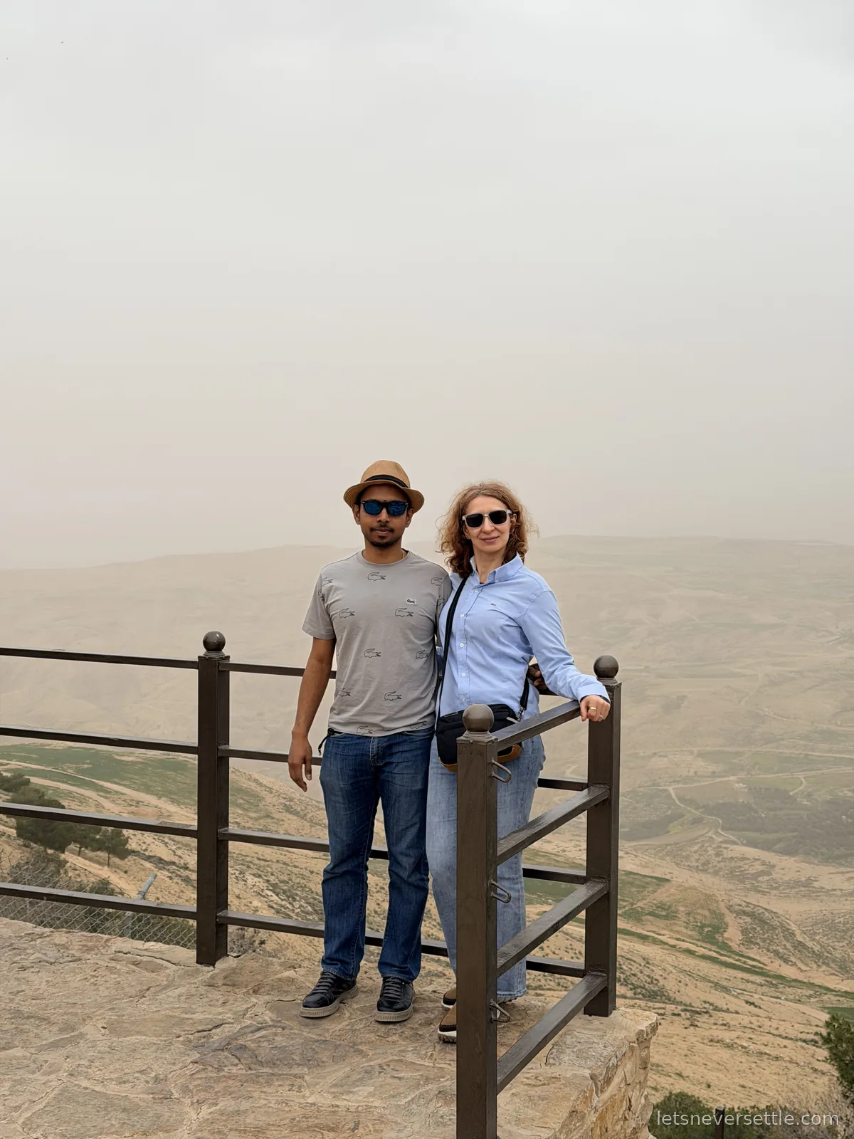 Inga and Dileep at the railing on the Mount Nebo summit, the hazy Jordan Valley dropping away behind them