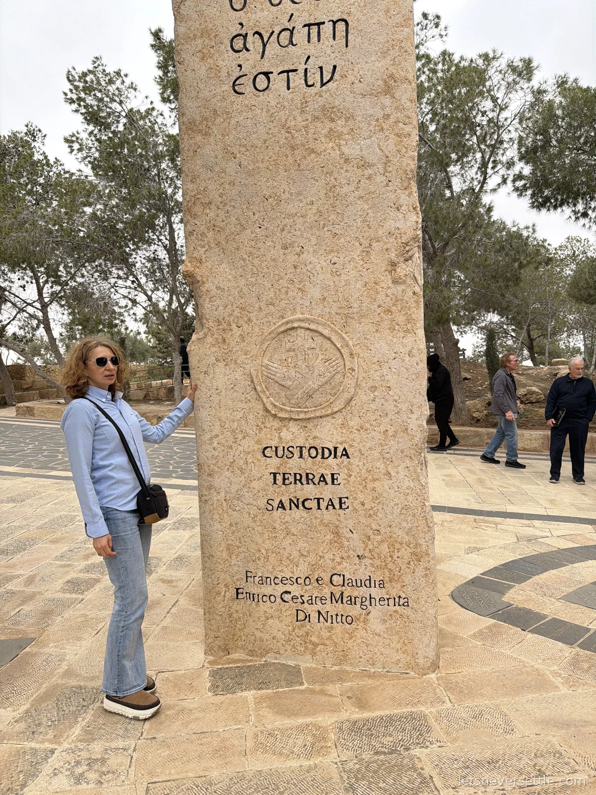 Inga resting a hand on a tall carved stone pillar on Mount Nebo inscribed Custodia Terrae Sanctae, with Greek lettering above it
