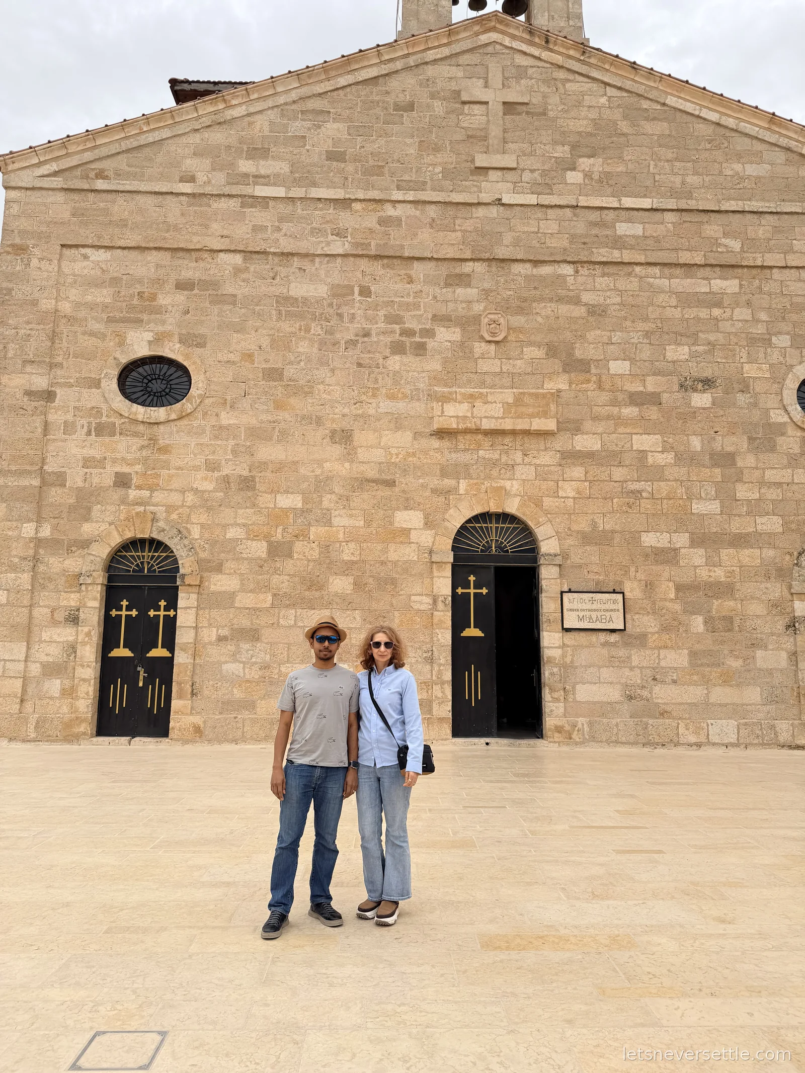 Inga and Dileep outside the stone facade of St George's Greek Orthodox Church in Madaba, with black and gold doors