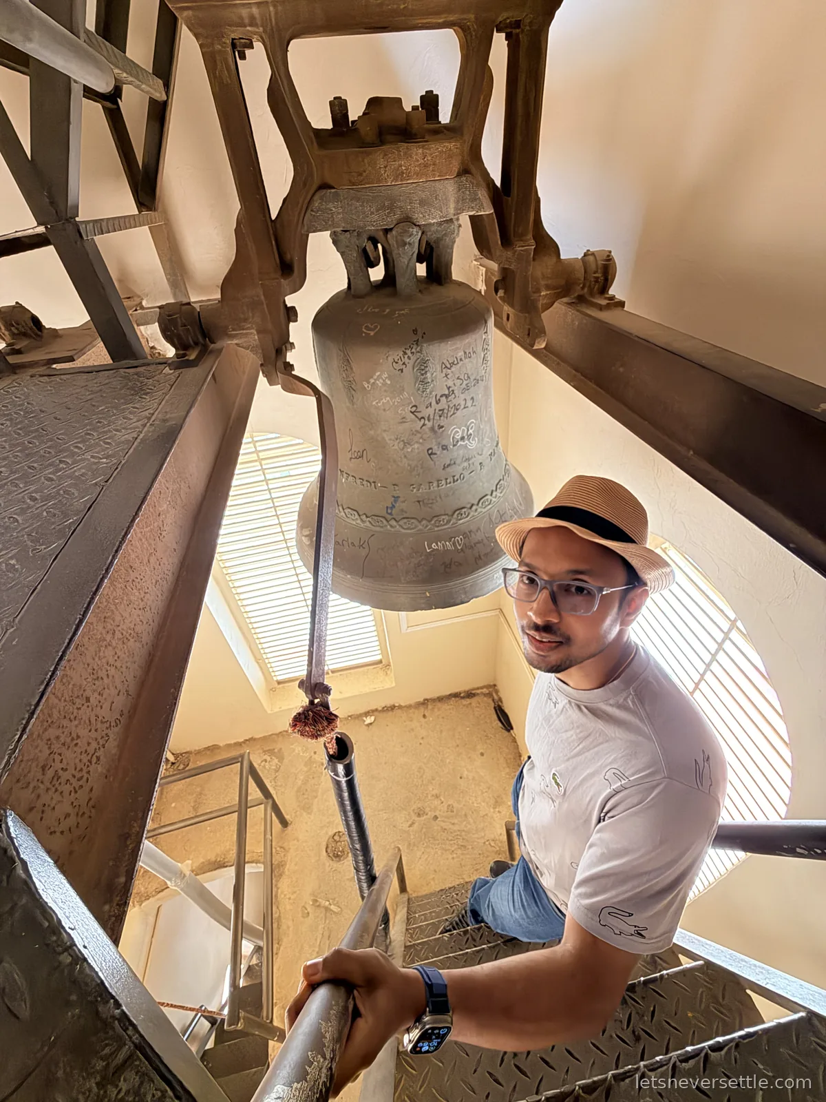Dileep on the metal staircase inside the bell tower of the Church of St John the Baptist in Madaba, beside a large bronze bell covered in scratched names