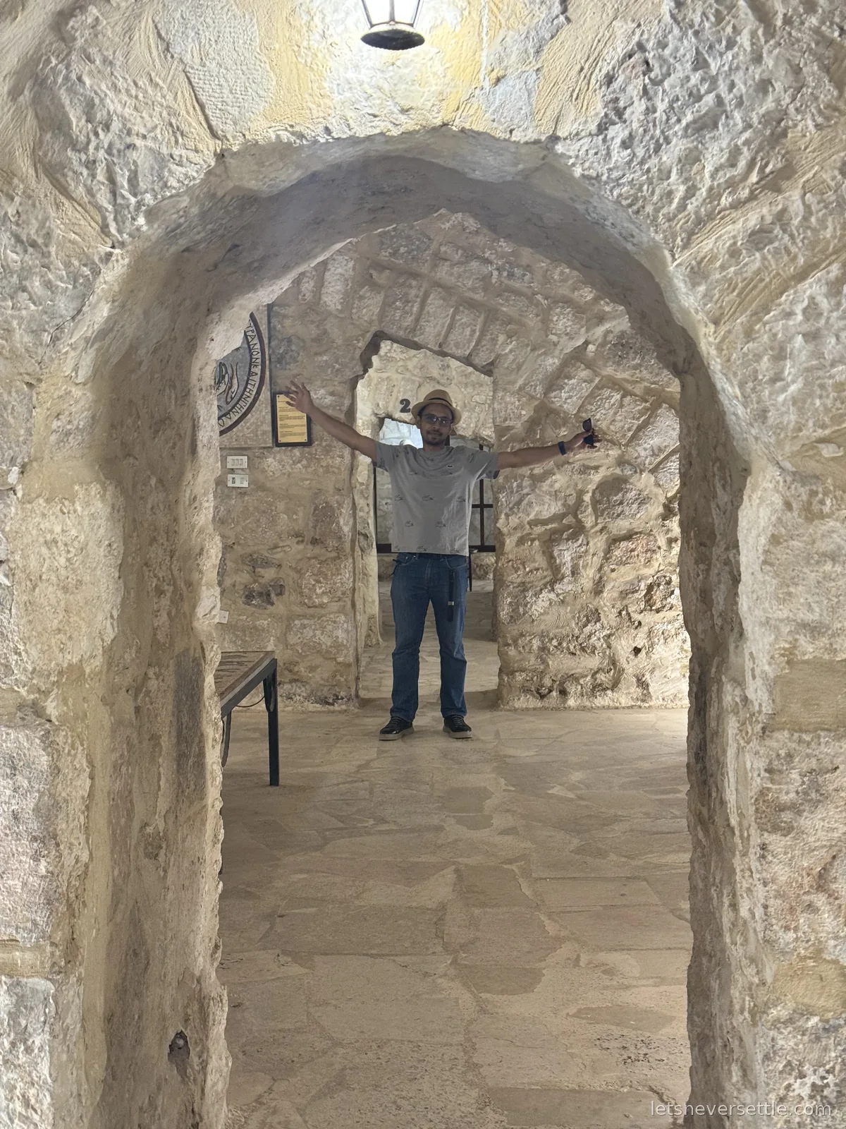 Dileep standing with arms outstretched in a narrow vaulted stone passage beneath the Church of St John the Baptist in Madaba, a lantern hanging from the arch