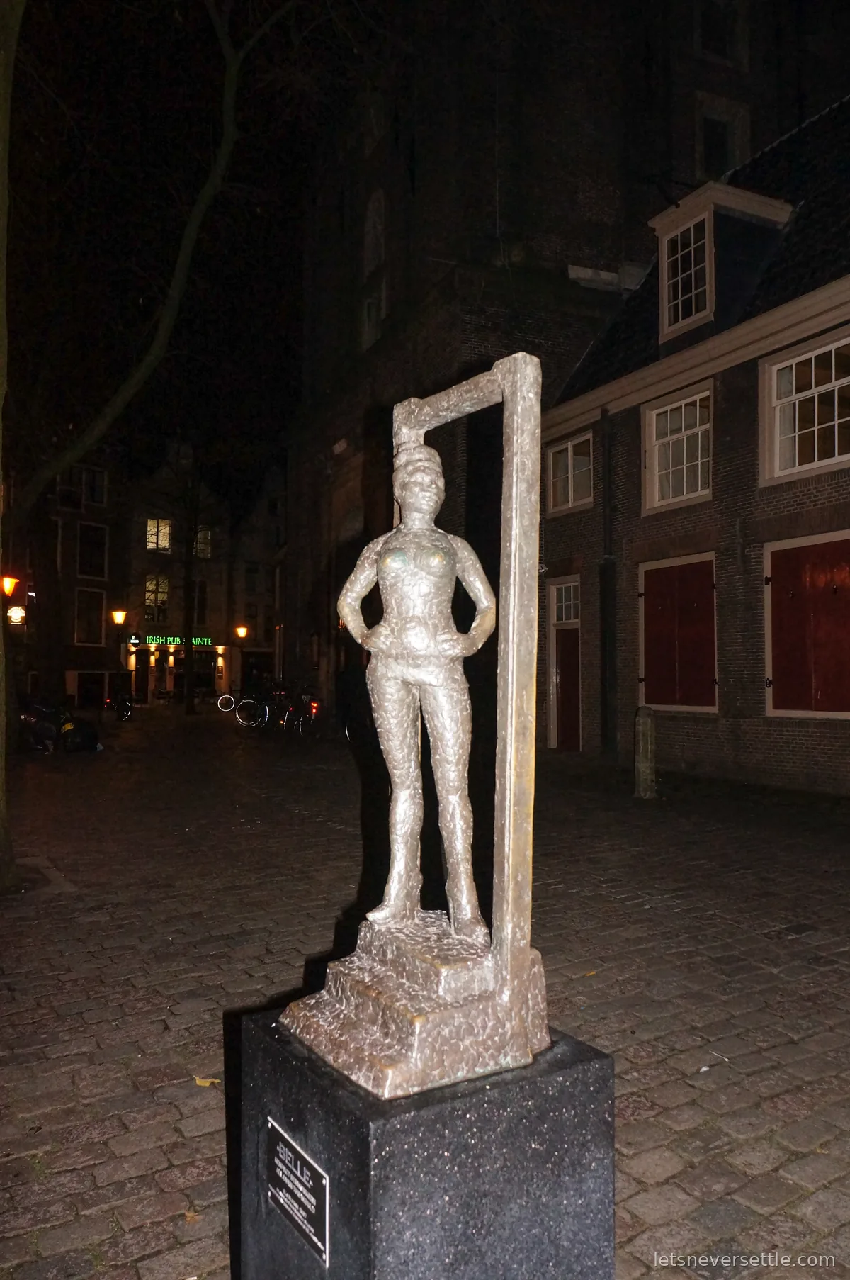 Belle, a bronze statue of a woman standing tall in a doorframe, on a dark cobbled square with red-shuttered windows behind