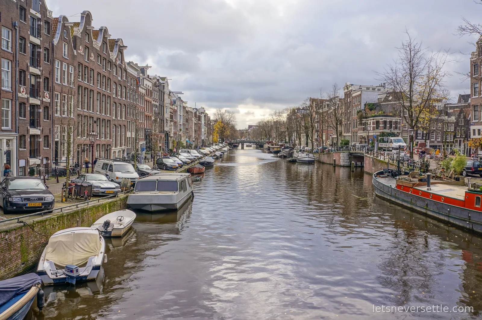 An Amsterdam canal lined with brick gabled houses, parked cars, small boats and an orange houseboat on grey water