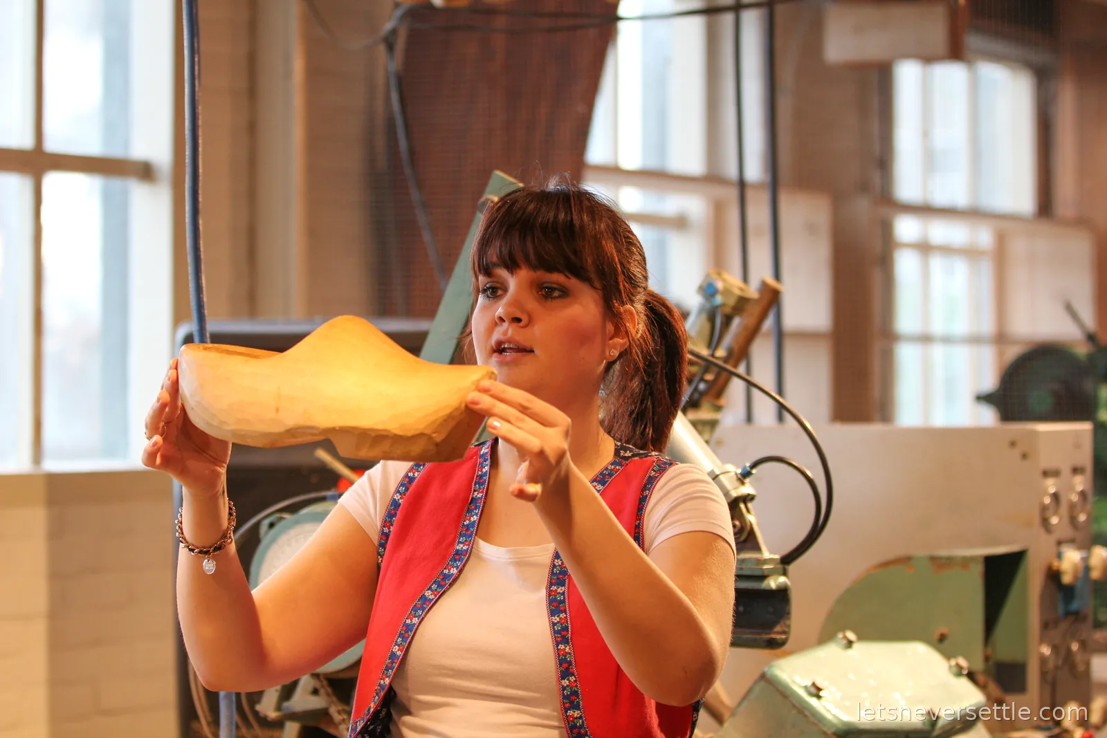 A demonstrator in a red traditional Dutch vest holding up a freshly carved pale wooden clog in a workshop