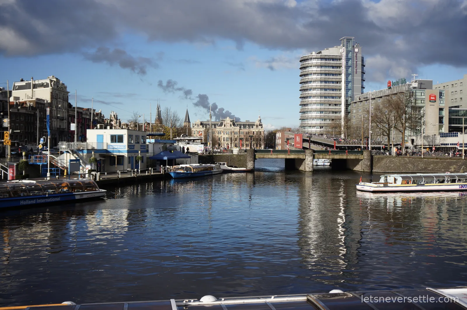 Canal cruise boats moored near Amsterdam Centraal under a blue winter sky, a round modern tower and hotels behind