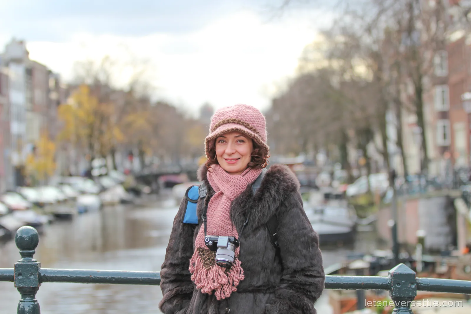 Inga in a pink knitted hat and dark fur coat smiling on a canal bridge, moored boats blurred behind her