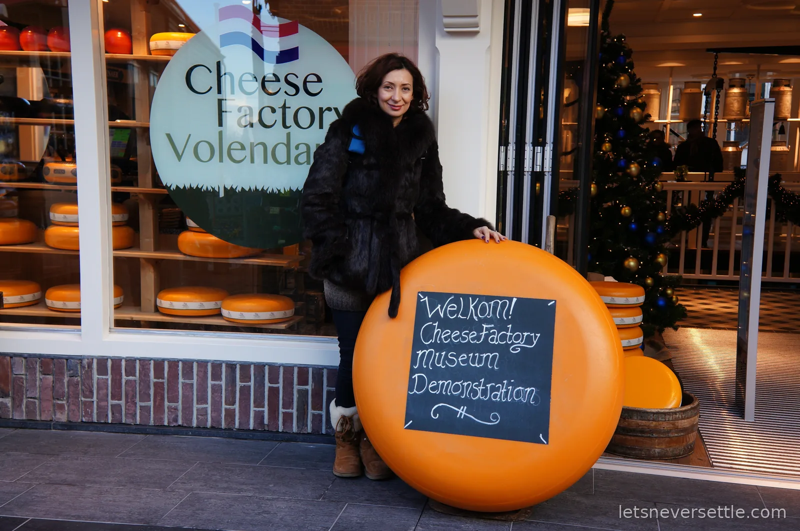 Inga in a fur coat holding a cheese wheel prop the size of a table, painted with Welkom Cheese Factory Museum Demonstration
