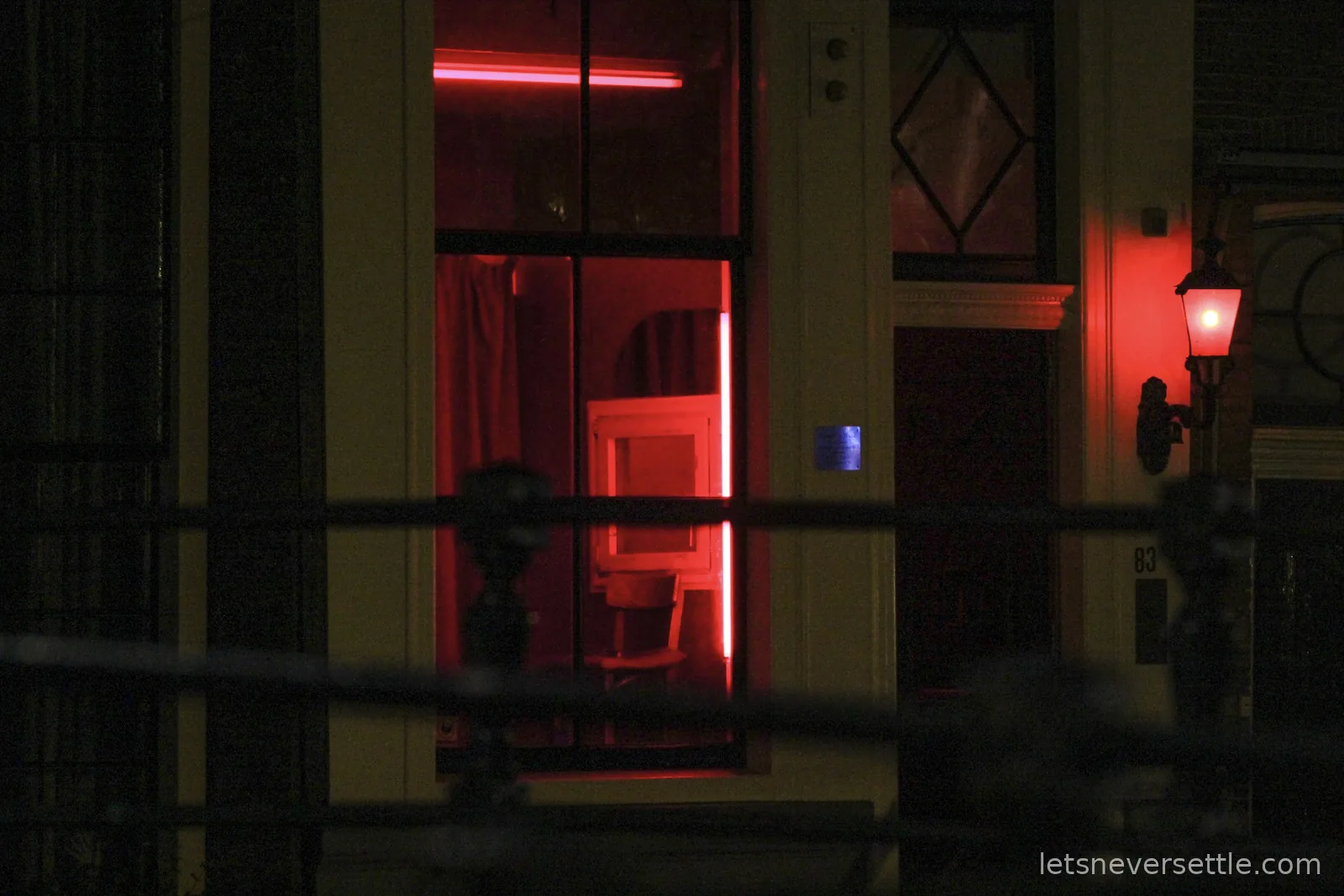 A window glowing deep red at night with an empty chair inside, next to a door with a red lantern and the number 83