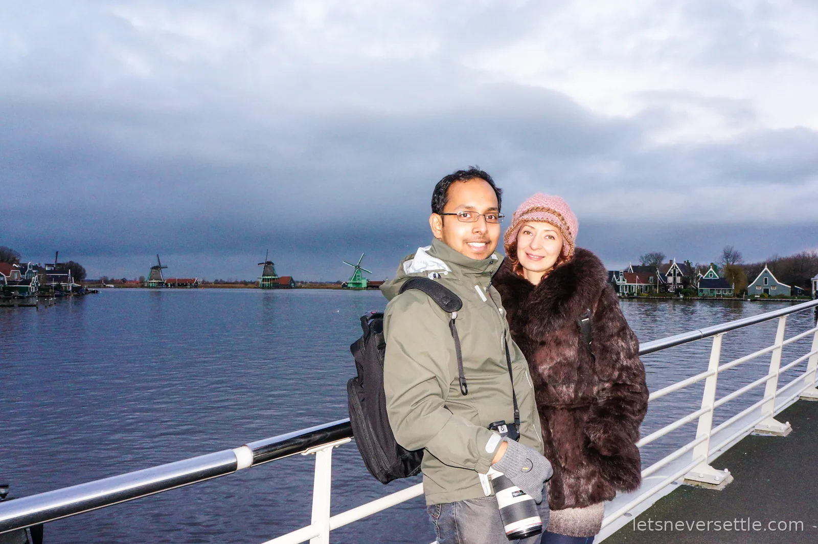Inga and Dileep standing together by a white railing with a row of Zaanse Schans windmills and green wooden houses across the water