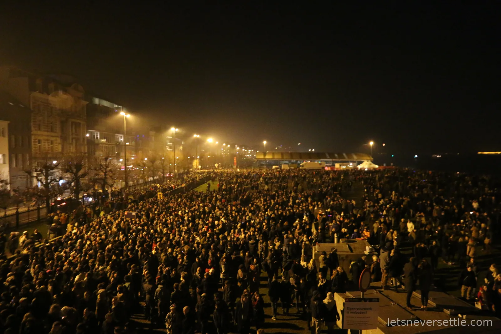 Huge crowd of people filling the Scheldt quays in Antwerp at night, under street lights, with a Dutch sign in the foreground banning private fireworks