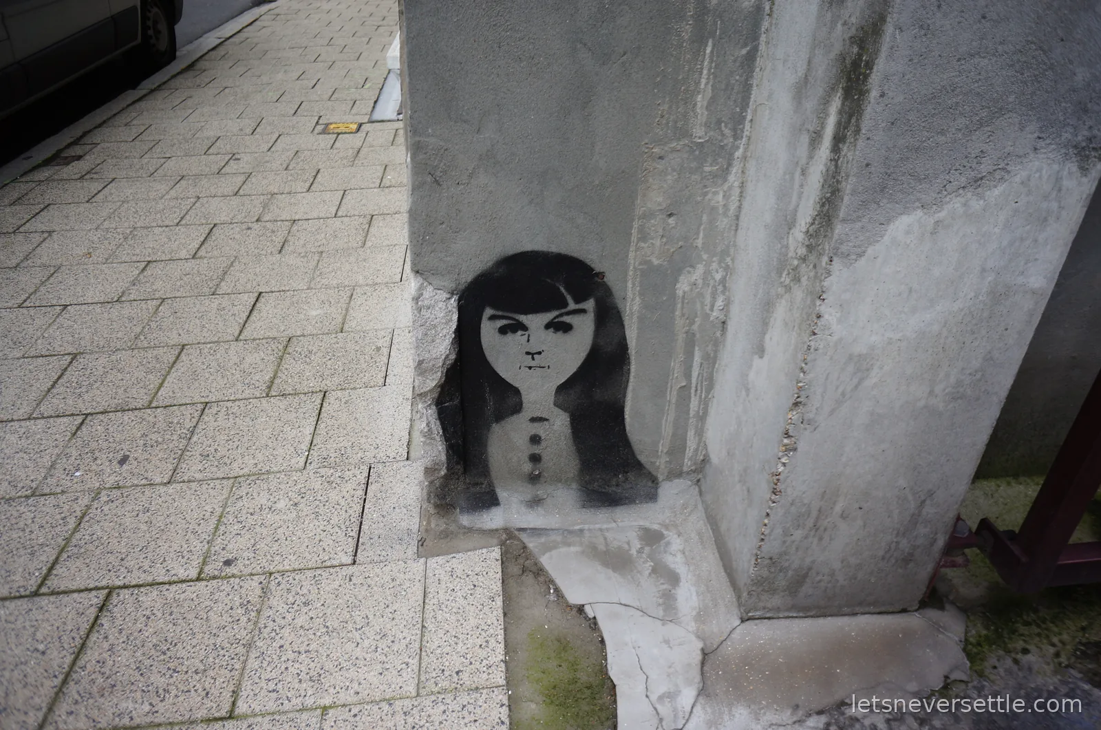 Black stencil graffiti of a girl with long dark hair, painted into a broken patch of plaster on a grey wall corner beside a paved pavement