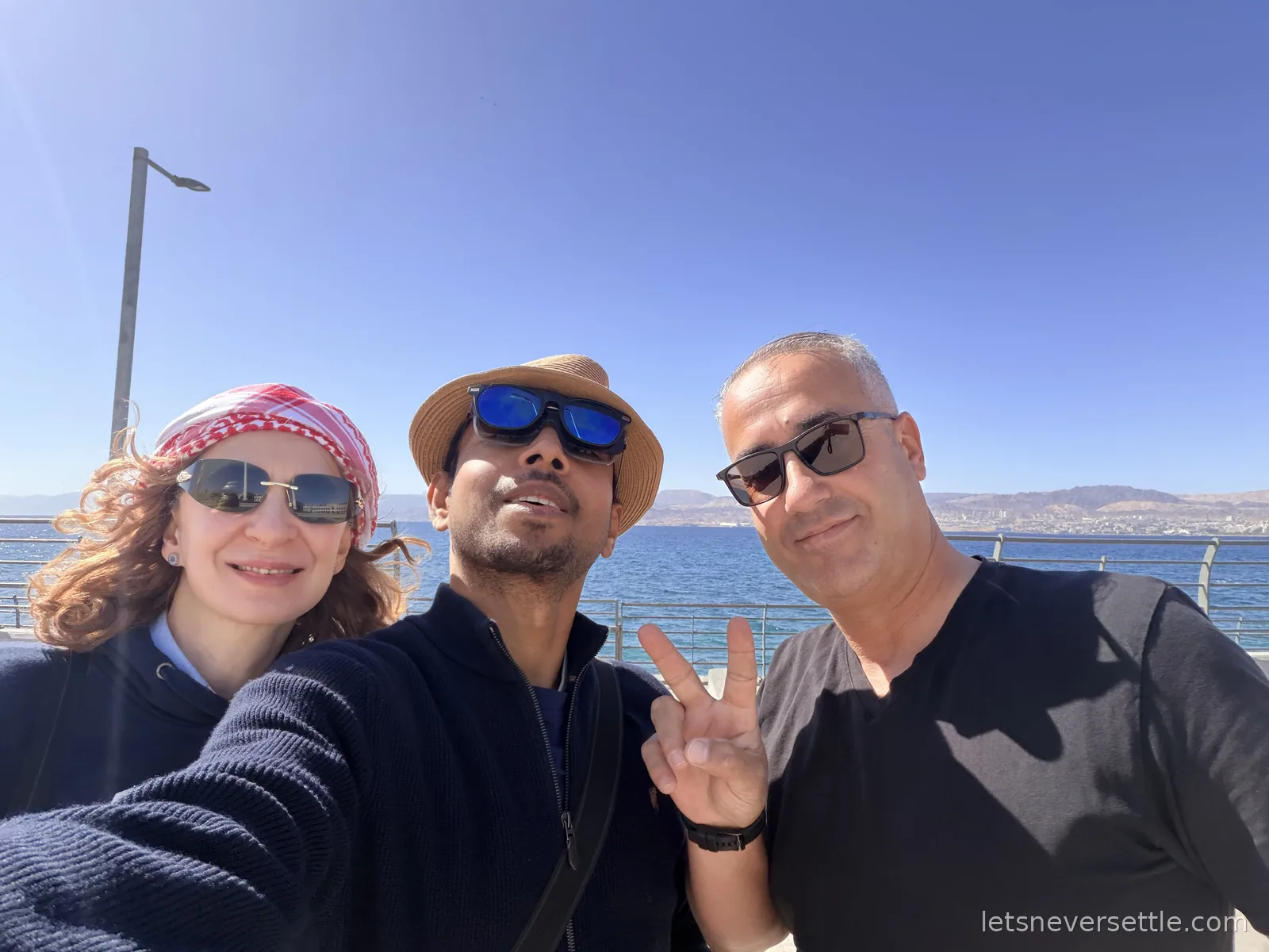 Inga, Dileep and a third member of their group taking a selfie on the Aqaba waterfront, the Red Sea and the hills of the far shore behind them