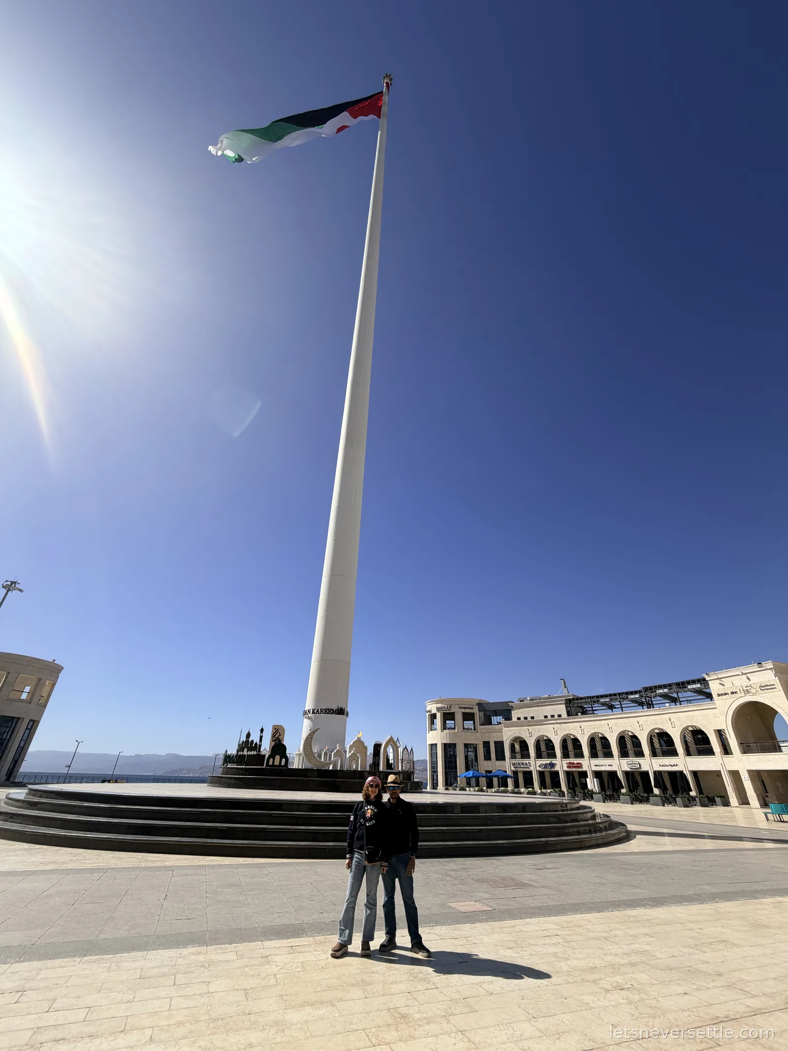Inga and Dileep standing beneath Aqaba's giant flagpole, the huge flag flying against a deep blue sky with Ramadan decorations on the plaza