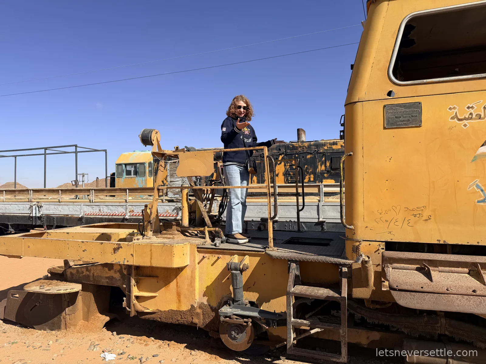Inga standing on the running board of a weathered yellow locomotive parked on the Hejaz Railway line in Wadi Rum, open desert behind