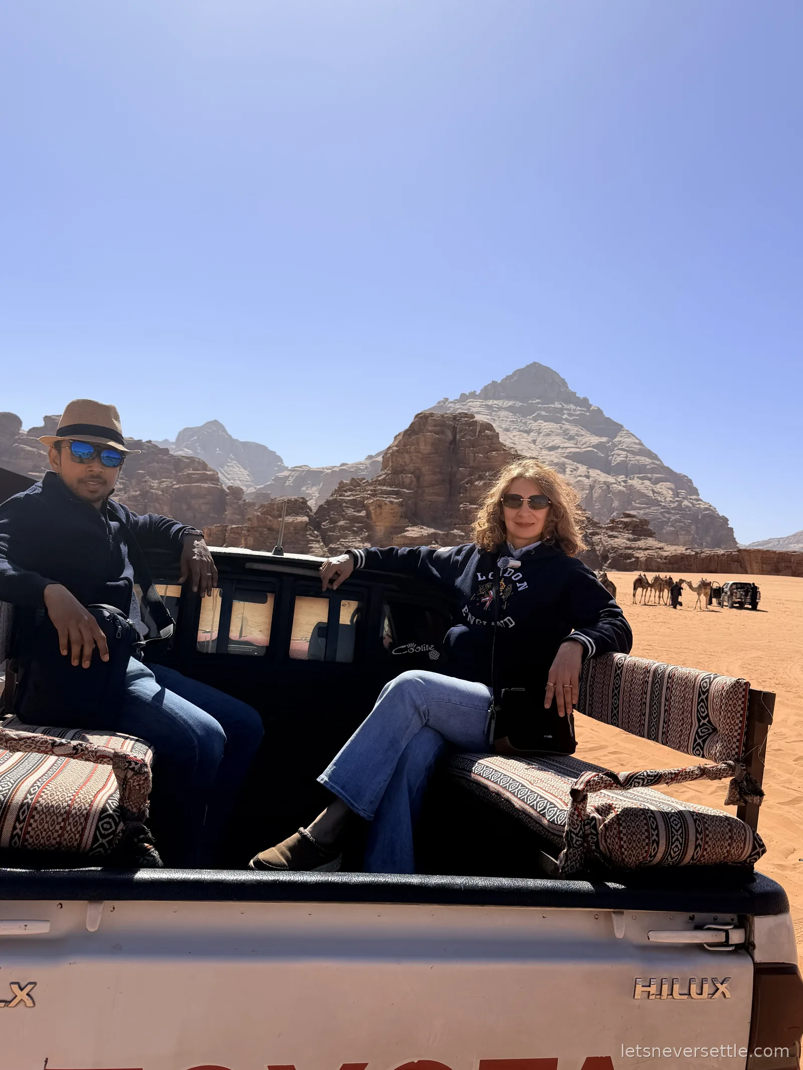 Inga and Dileep sitting on patterned cushions in the open back of a Toyota Hilux in Wadi Rum, with camels and sandstone cliffs behind