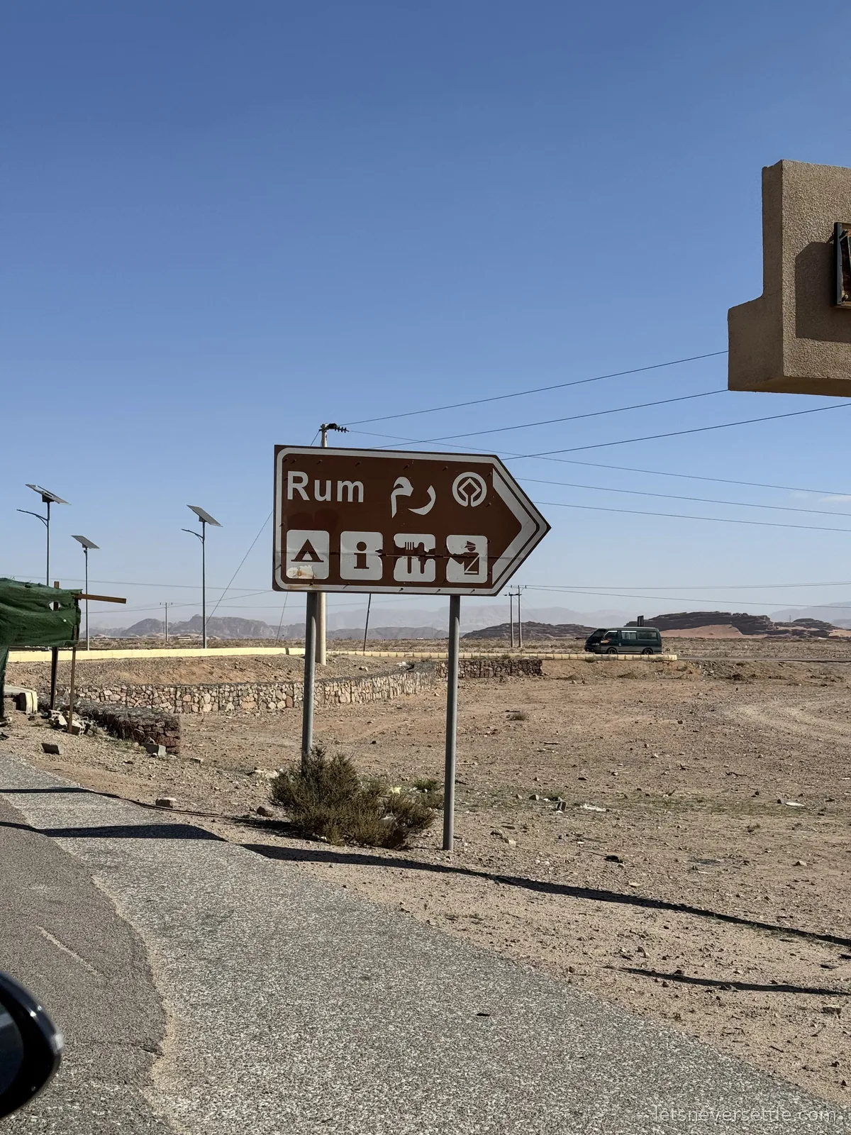 A brown road sign reading Rum in English and Arabic, pointing off a desert highway towards the Wadi Rum village