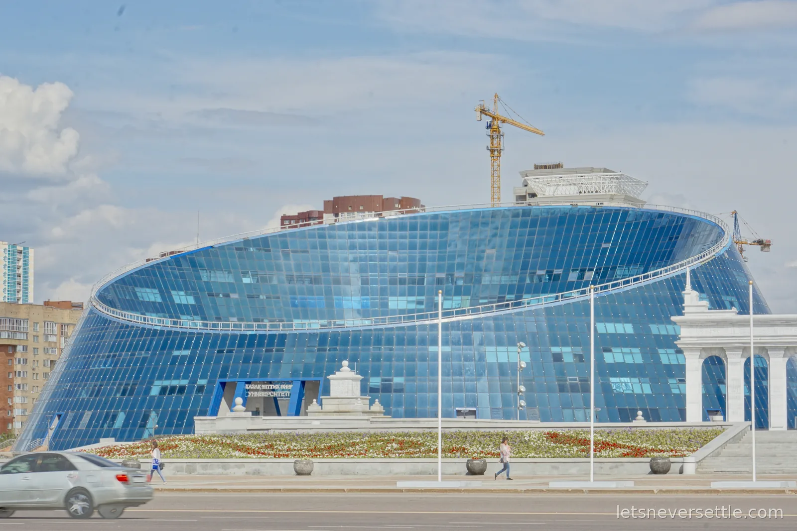 The Kazakh National University of Arts, a tilted ellipse of blue glass, with a construction crane rising behind