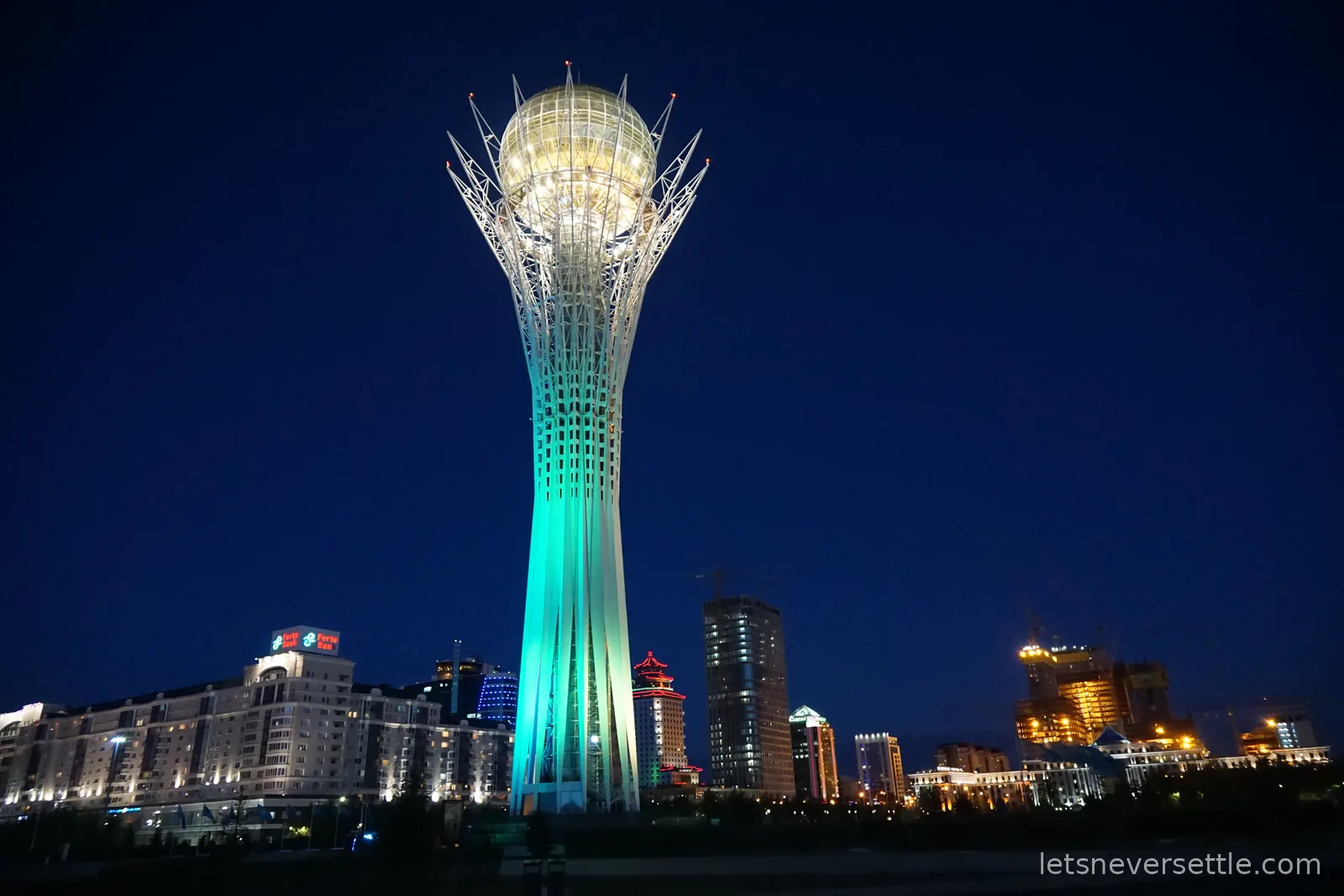 The Bayterek tower at night, its white latticed column lit turquoise beneath a golden glass sphere, lit towers of central Astana around it