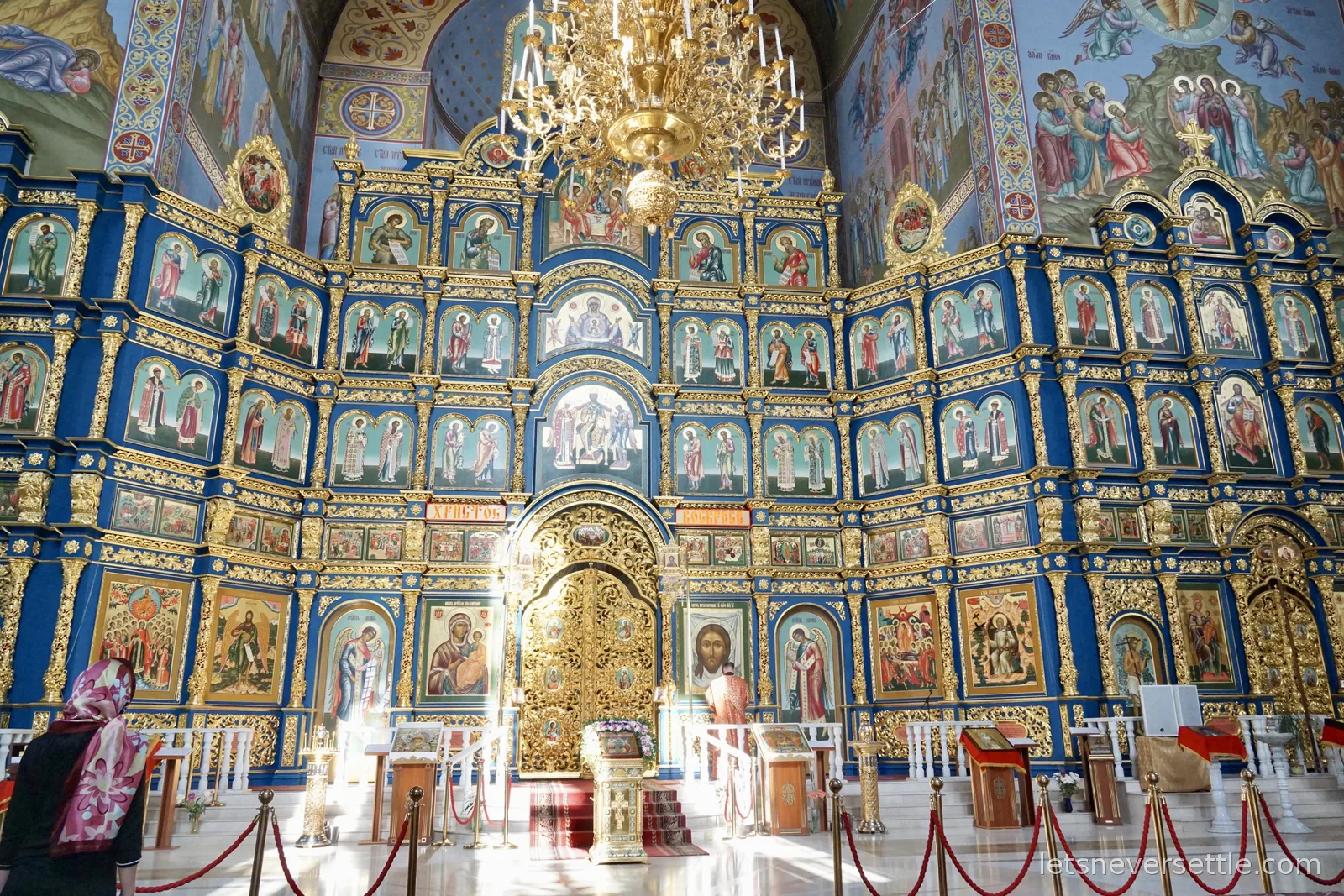A cathedral iconostasis in deep blue and gold rising toward painted vaults, rows of icons around a gilded central door, a chandelier above