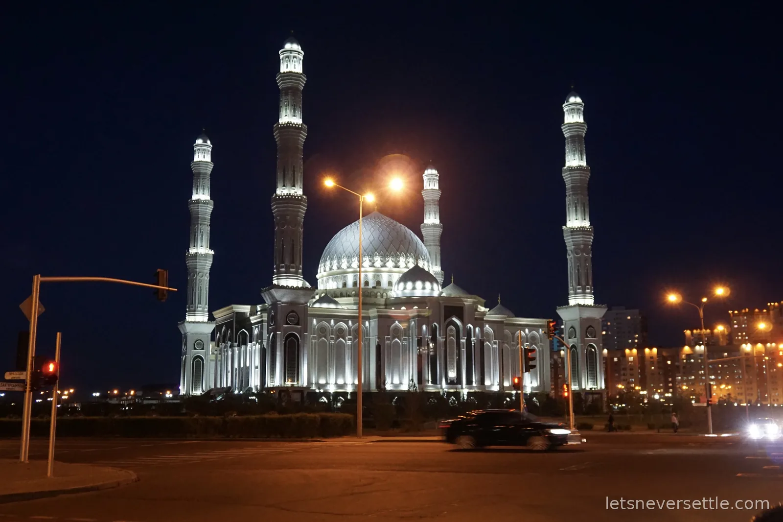 The white Hazrat Sultan Mosque floodlit at night, four slender minarets around a patterned central dome, a car passing in front