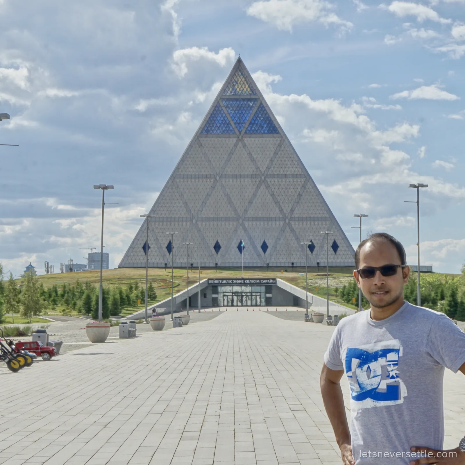 Dileep in sunglasses in front of the Palace of Peace and Reconciliation, a grey stone pyramid with stained glass panels at its apex