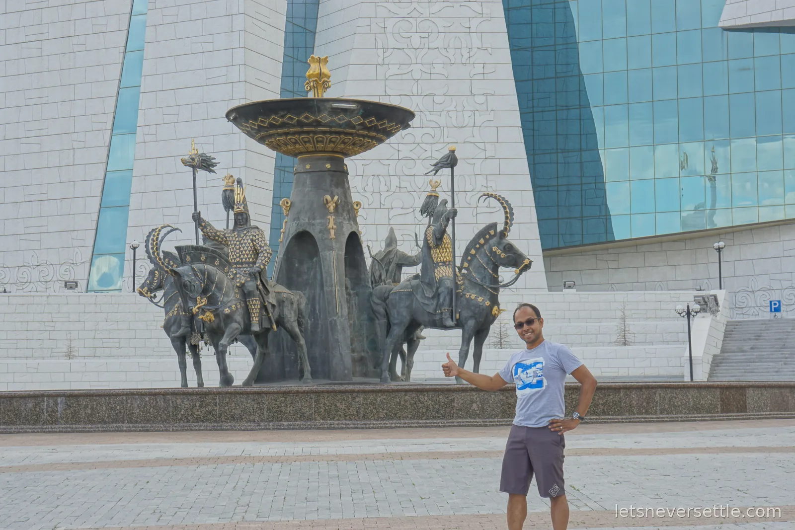 Dileep giving a thumbs up beside a fountain of Saka horsemen statues in gilded armour, the white stone and glass facade of the National Museum behind