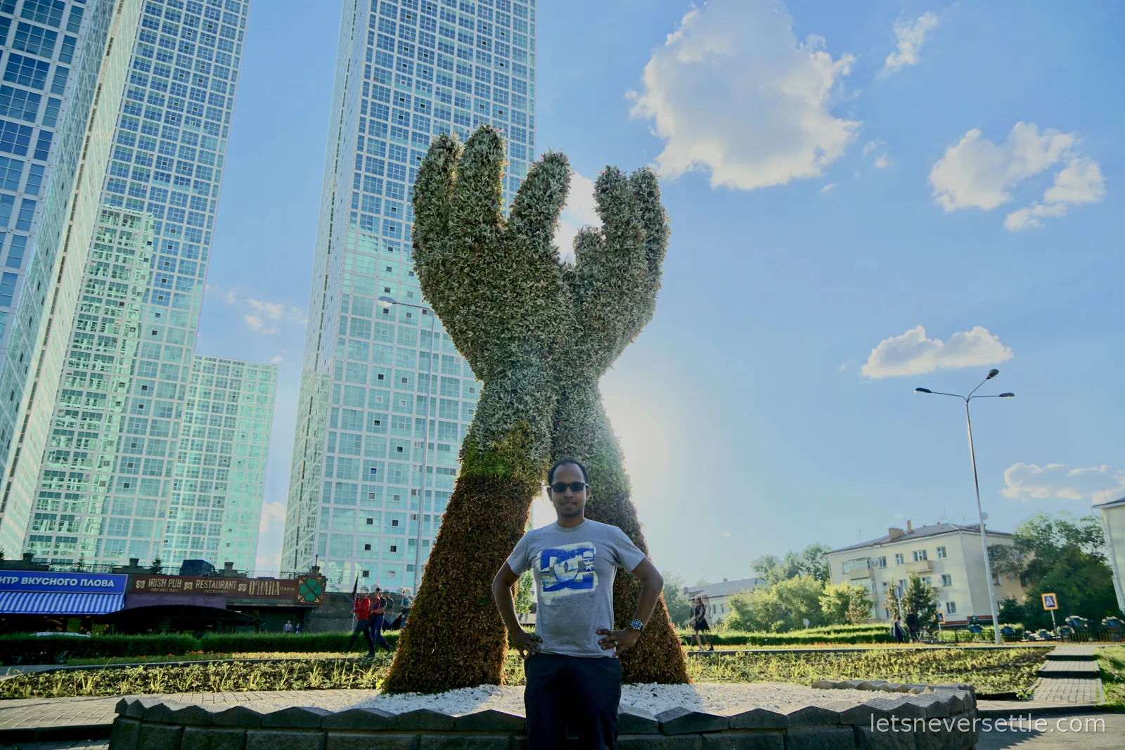 Dileep standing in front of a giant topiary sculpture of two raised hands, glass apartment towers filling the sky behind